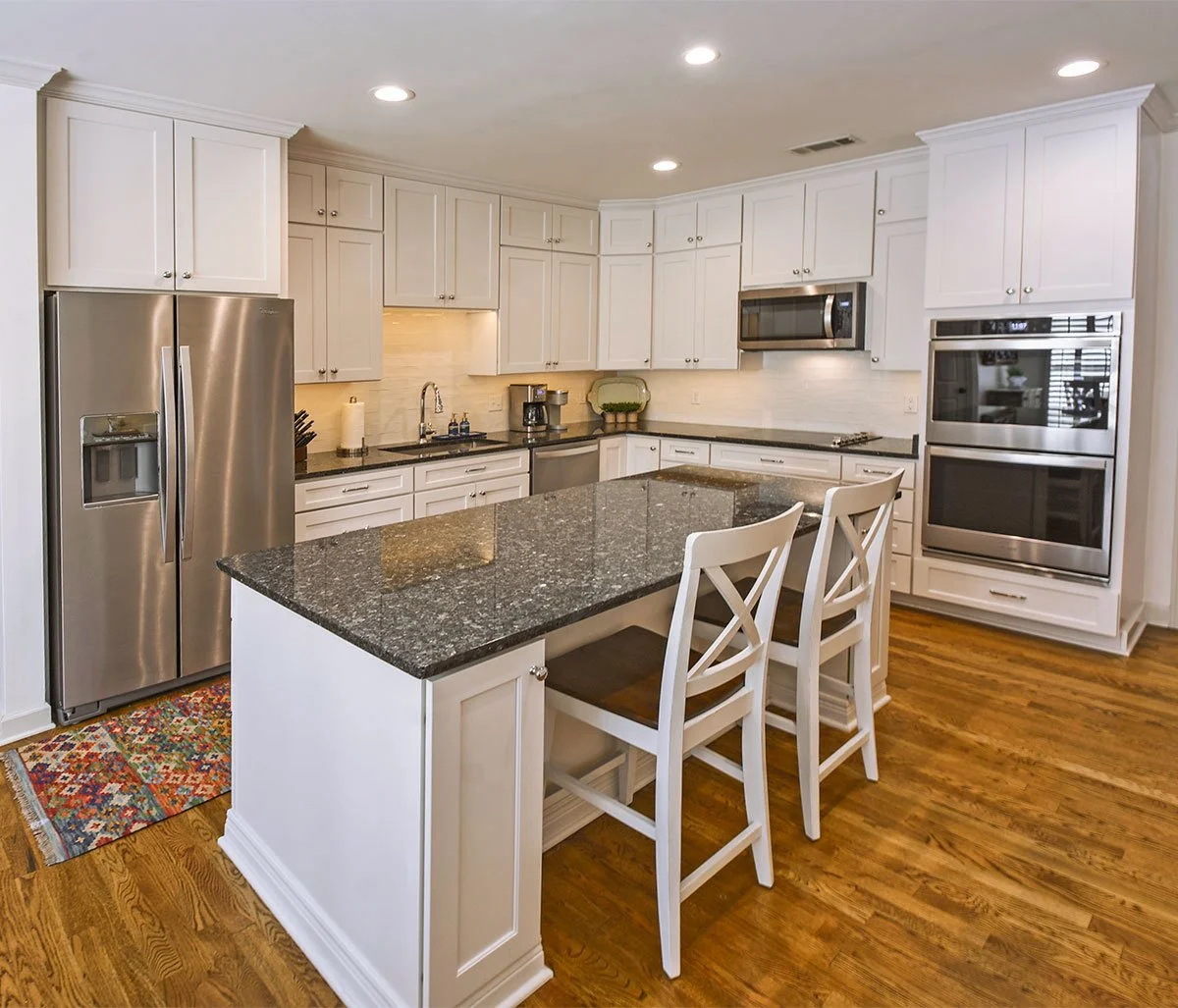 A modern kitchen with white cabinets, stainless steel appliances, a granite island, and hardwood flooring.
