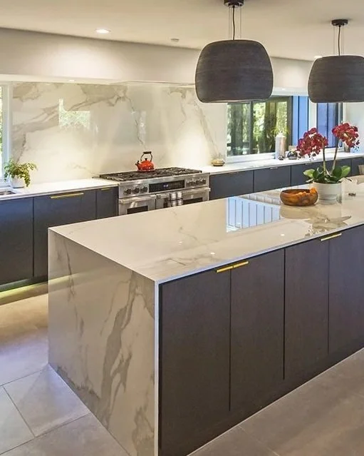Modern kitchen with dark cabinets, white marble island, marble backsplash, two pendant lights, and a window with red flowers.