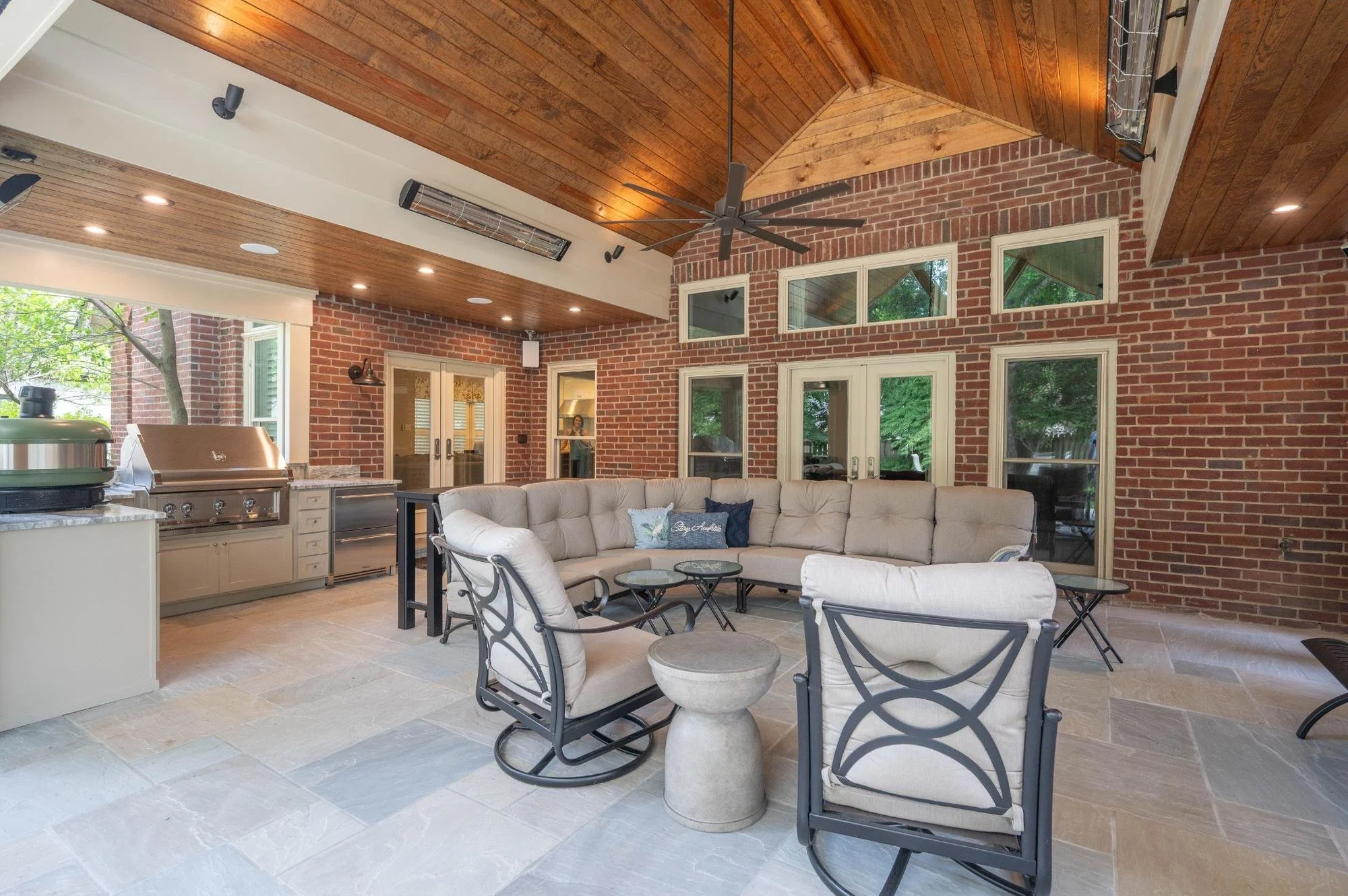A covered outdoor patio with a brick wall, wooden ceiling, ceiling fan, and glass doors leading inside. The patio has white cushioned seating, black metal chairs, and small round tables, with a gas grill and outdoor heater visible.