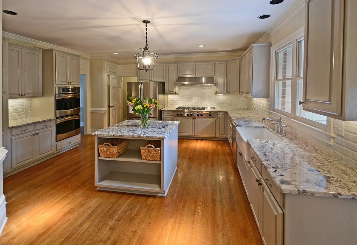 Modern kitchen with white cabinets, granite countertops, hardwood floors, a central island with a vase of flowers, stainless steel appliances, and large windows.
