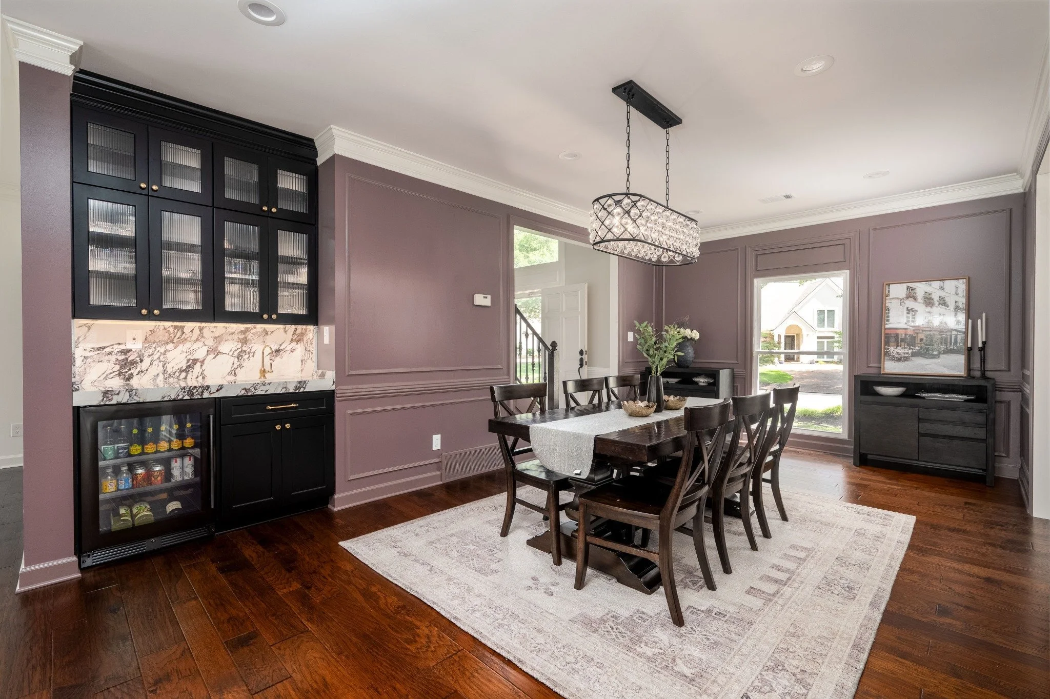 Dining room with a wooden table, black chairs, a chandelier, purple walls with decorative molding, a floral arrangement, and a large window showing a residential street outside.
