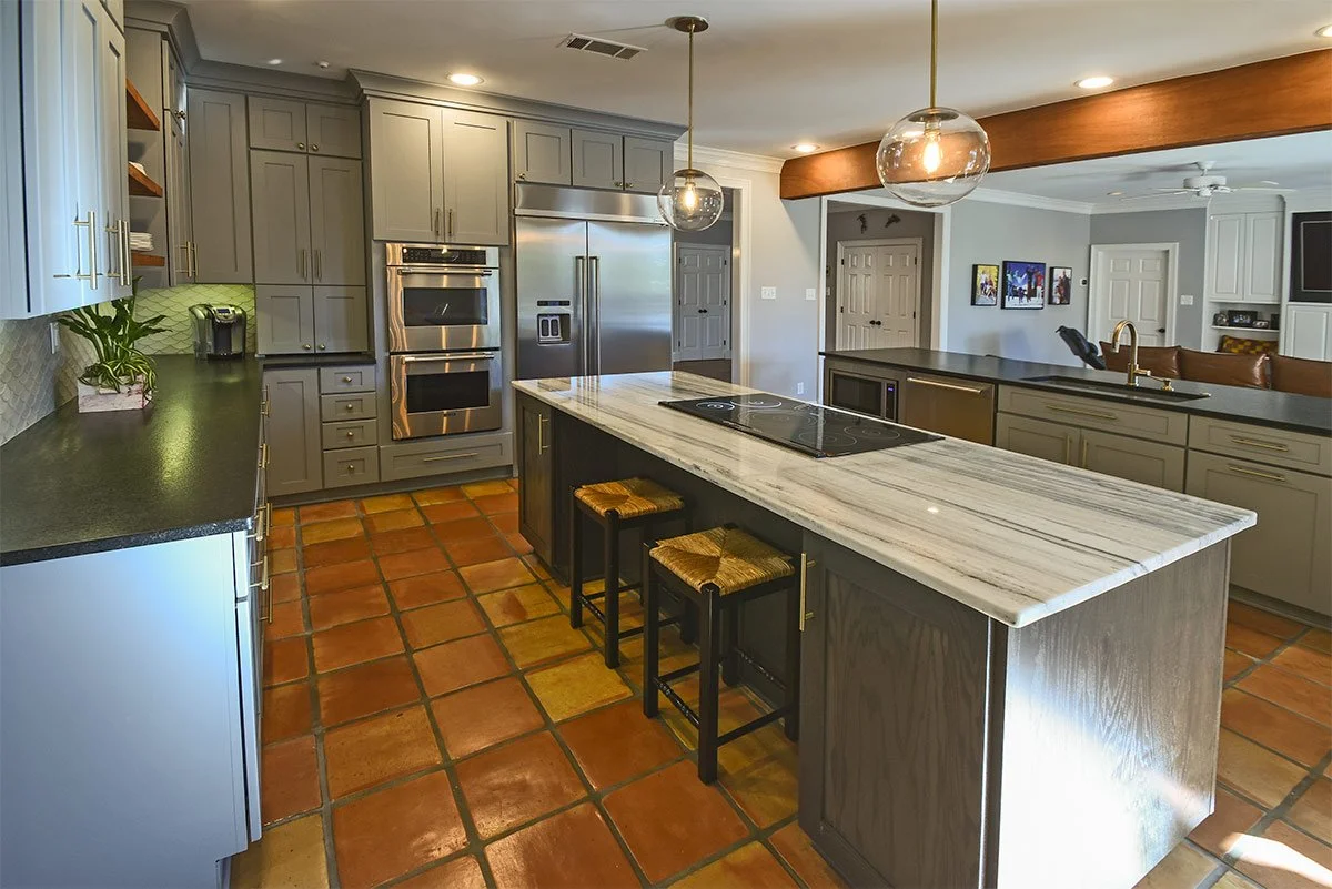 Kitchen with gray cabinets, stainless steel appliances, a large island with a marble countertop, terracotta tile flooring, and two pendant lights hanging above the island.