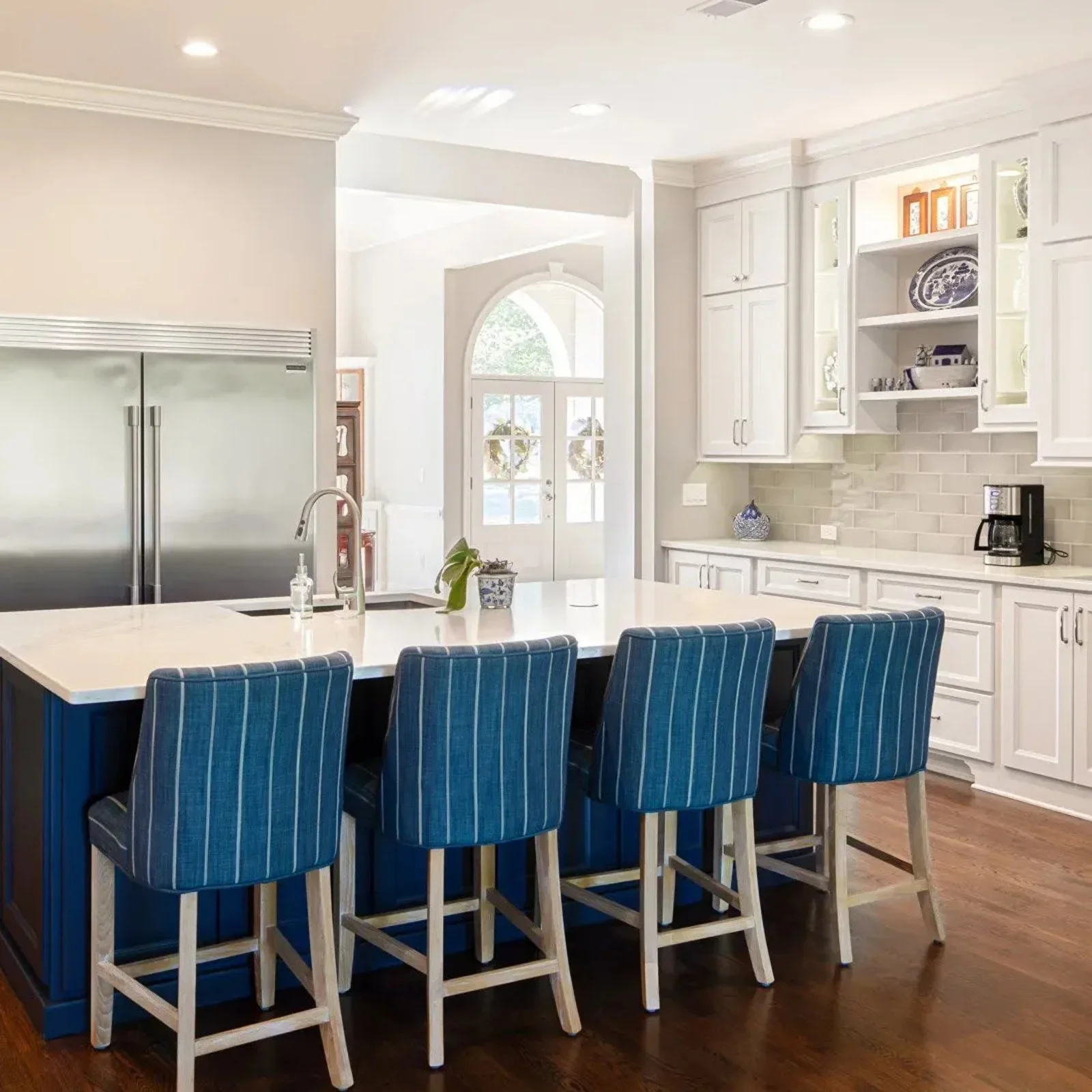 Kitchen with white cabinetry, a large white island with blue paneling, and five blue and white striped chairs. There is a stainless steel refrigerator to the left, and a doorway with an arched window leading outside. The countertop has a sink, a small plant, a soap dispenser, and a coffee maker. The room has recessed lighting and hardwood flooring.