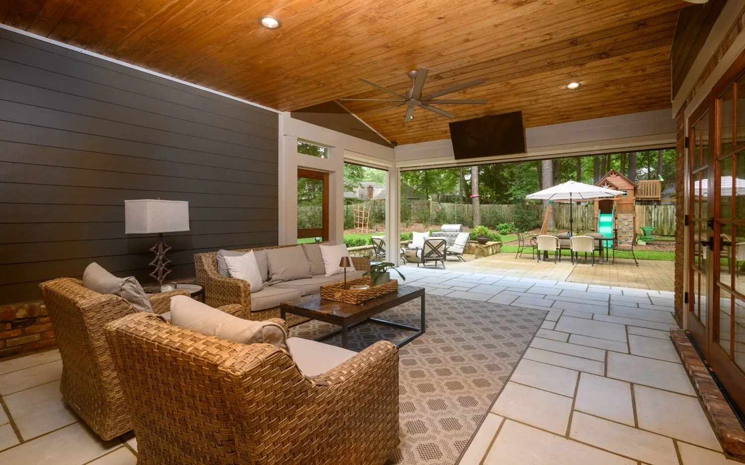 Covered porch area with wicker seating and a rug, opening to a backyard with outdoor furniture, umbrella, playhouse, and slide.