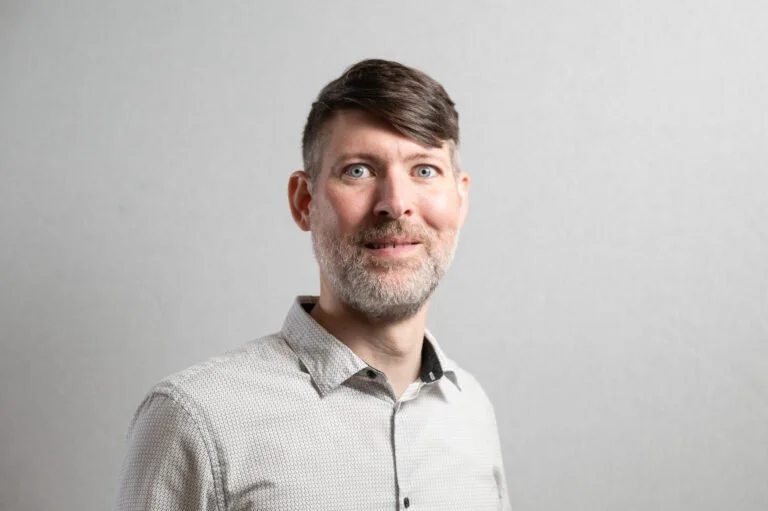 Portrait of a man with gray hair and beard, wearing a light gray shirt, standing against a plain gray background.