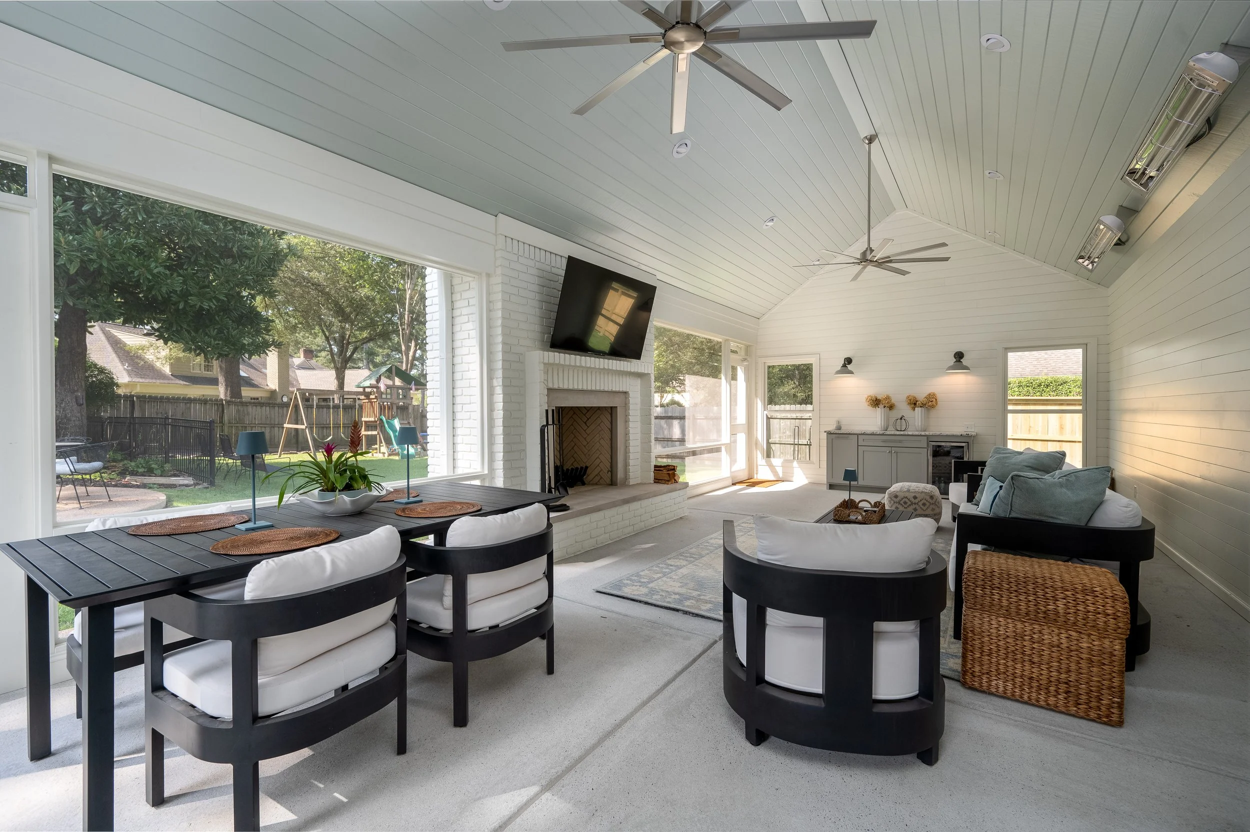 Covered patio with large windows, a ceiling fan, a fireplace, a dining table with chairs, and a seating area with sofas and a woven chair. Outside, there is a backyard with trees, a wooden fence, a swing set, and outdoor furniture.