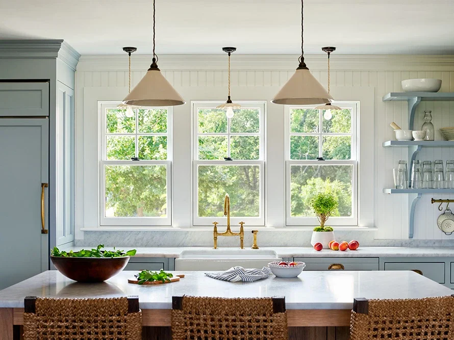 Bright kitchen with three large windows, white paneled walls, marble countertop, brass faucet, open shelves with dishes and glasses, and three pendant lights.