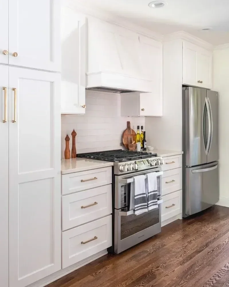 A modern kitchen with white cabinets, stainless steel refrigerator, and a stainless steel oven with a gas cooktop. There are decorative items like wooden salt and pepper shakers, a round wooden cutting board, and bottles of oil on the countertop. The kitchen has dark hardwood flooring.