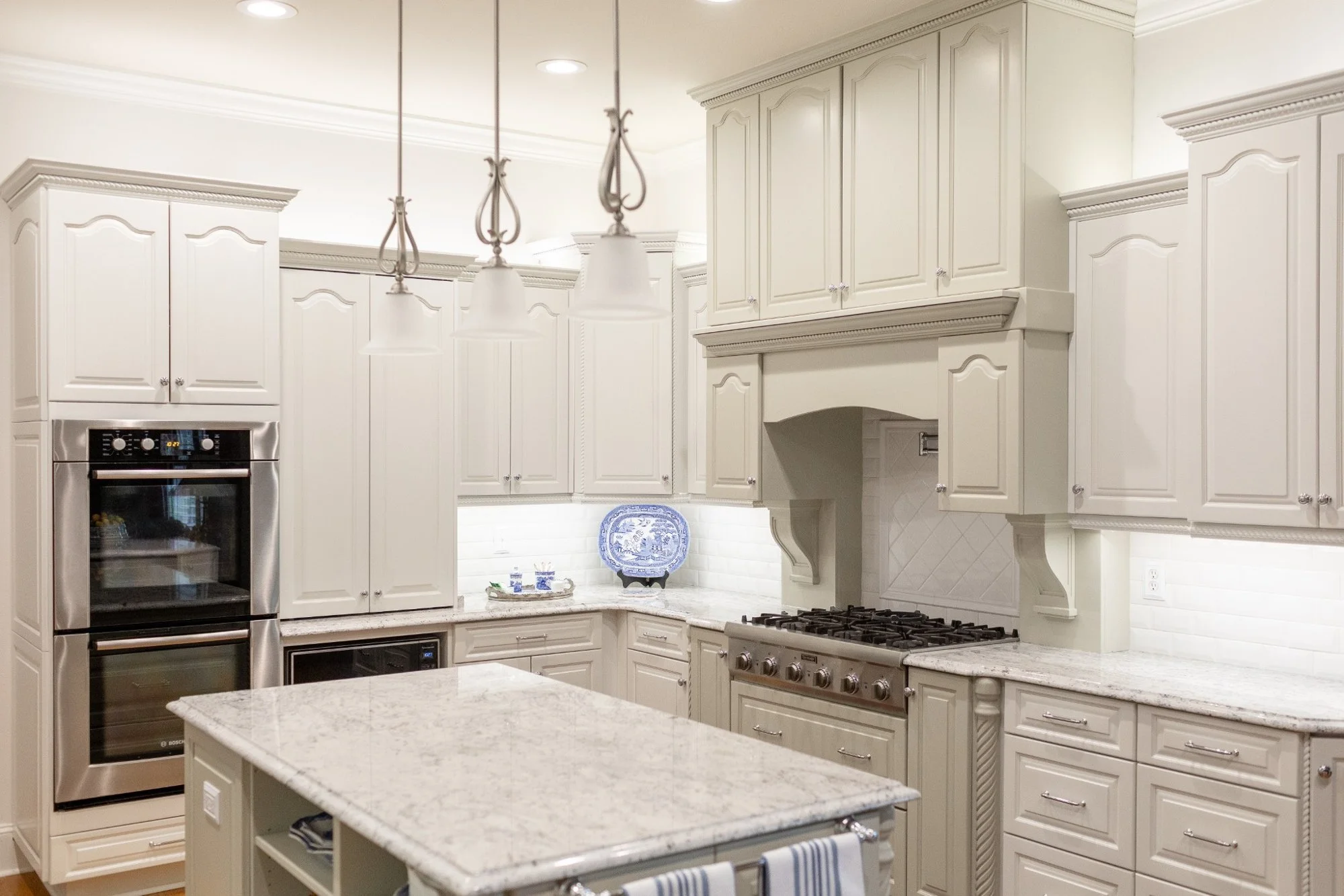 A spacious, elegant kitchen with white cabinets, a granite countertop island, a stainless steel oven, a gas cooktop, and pendant lighting over the island. Decorative molding and a display plate are visible.
