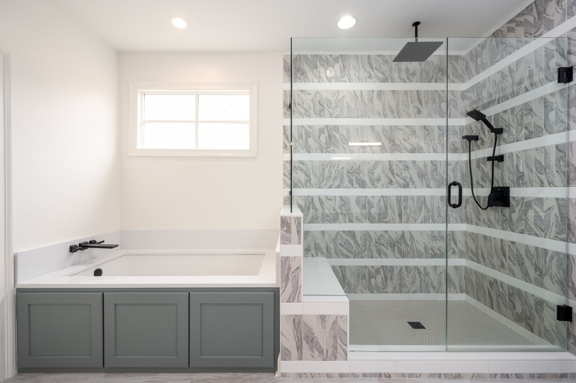 Modern bathroom with a separate walk-in shower area with gray and white patterned tiles, a rain shower head, and black fixtures. A window on the white wall provides natural light.