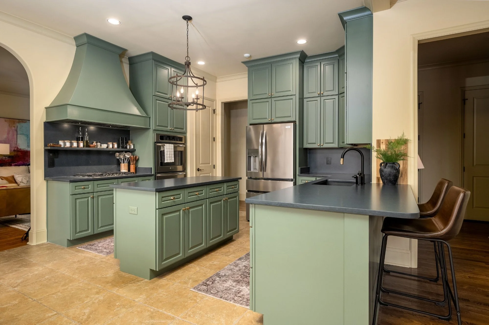 A kitchen with mint green cabinets, a black countertop, and a matching island. There is a stainless steel refrigerator, oven, and microwave. Over the island hangs a rustic black chandelier. There are brown barstools at the counter, a potted plant on the counter, and beige tile flooring.