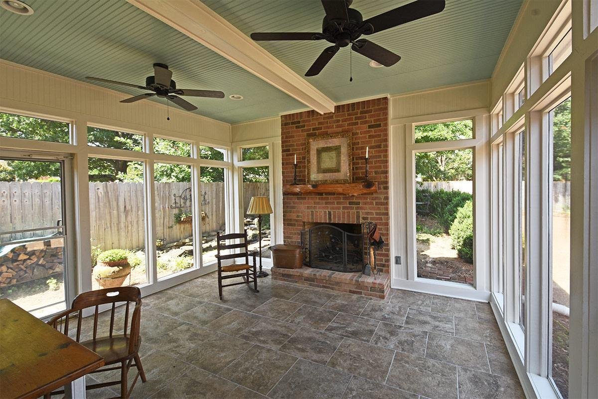 Sunroom with tile flooring, brick fireplace, ceiling fans, and large windows with a view of a backyard with trees and shrubs.