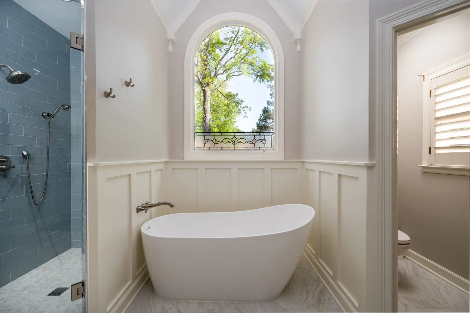 A luxurious bathroom with a freestanding bathtub positioned beneath a large arched window with decorative stained glass, and a separate walk-in shower with blue tiles. The room has white paneled walls, a small window with white shutters, and a view of green trees outside.
