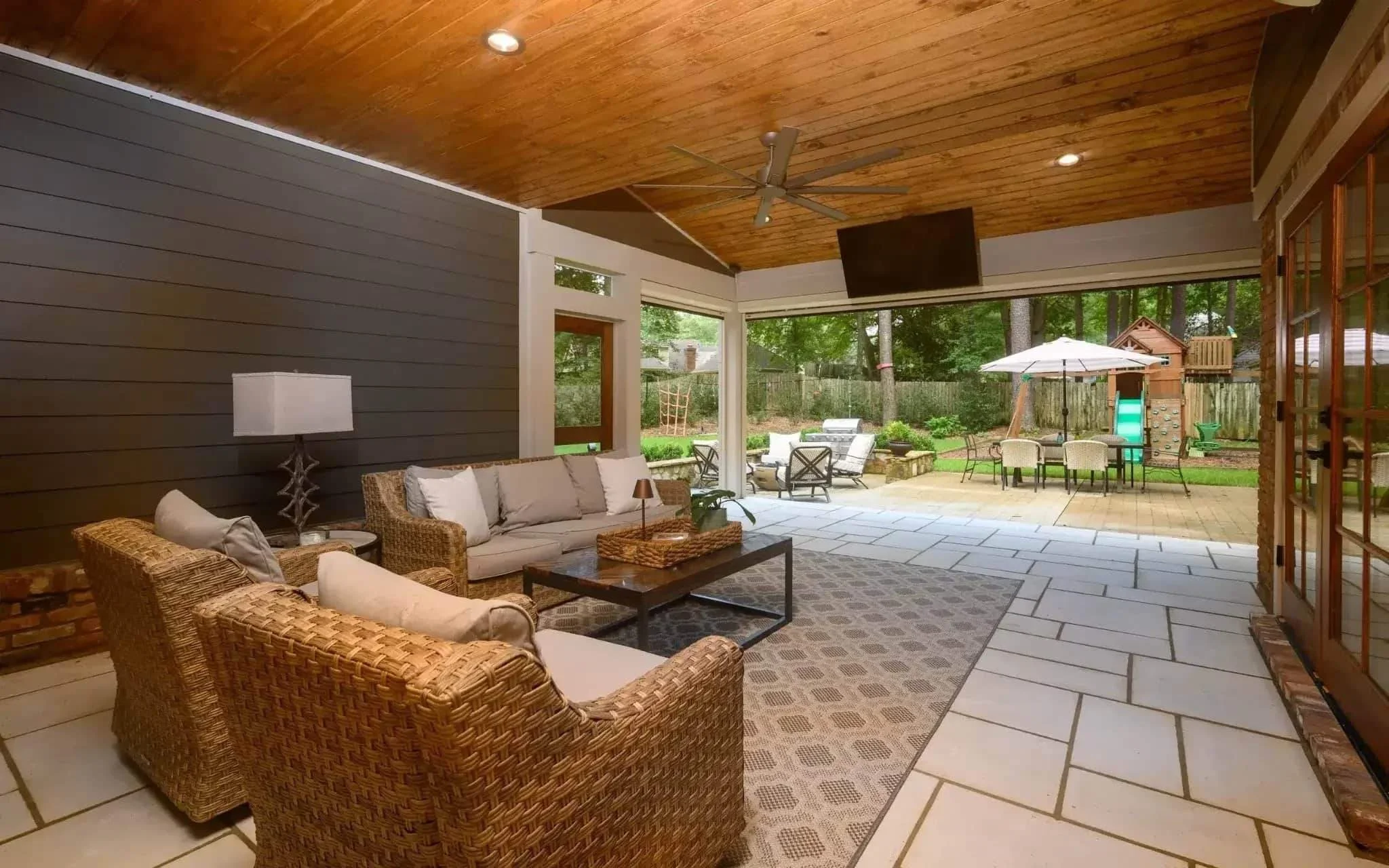 Custom covered outdoor living space with vaulted wood ceiling, comfortable lounge seating, ceiling fan, mounted TV, and open patio area designed for year-round entertaining in Germantown, TN.