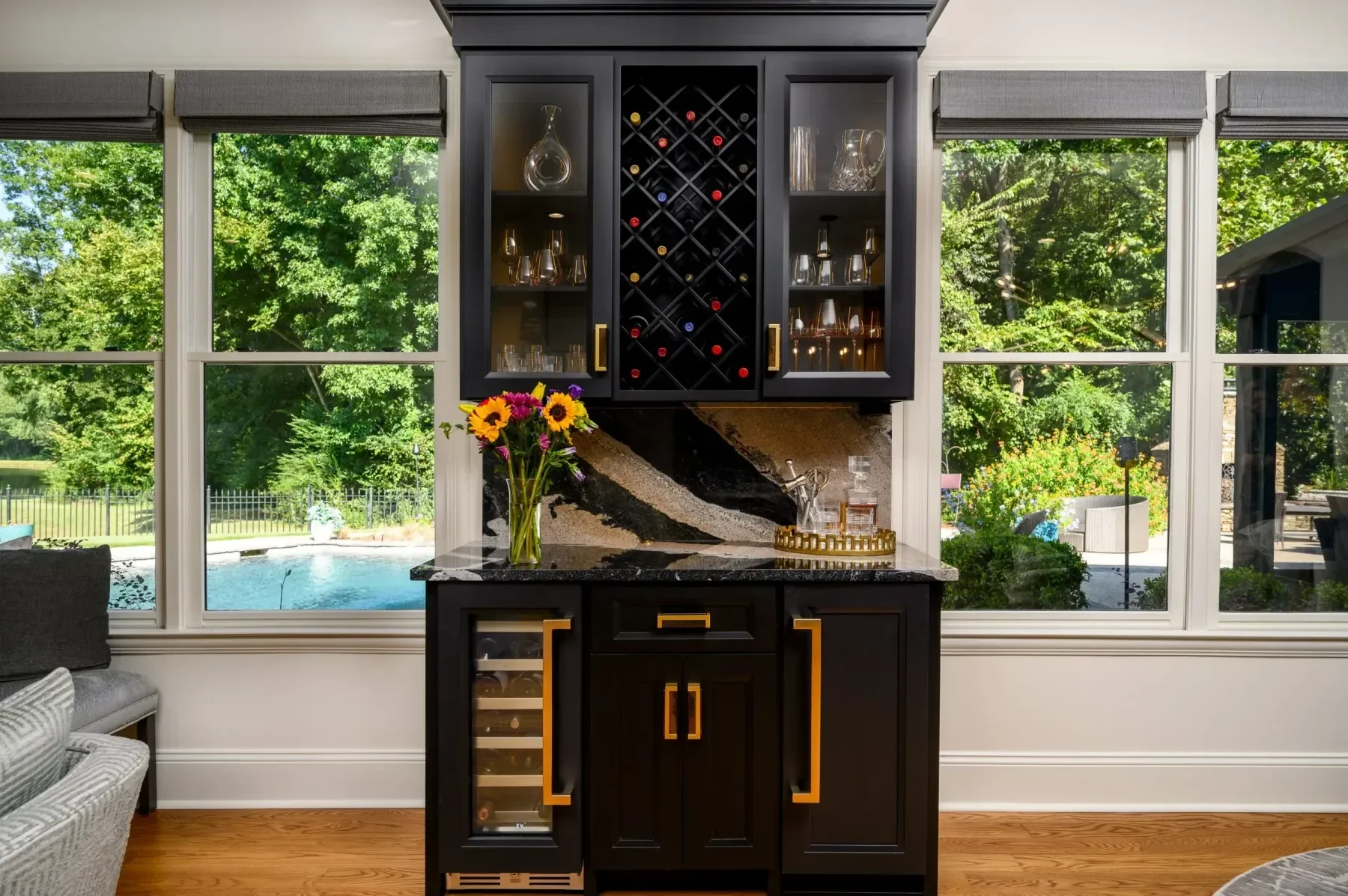 View of a black home bar with gold handles and accents, set against large windows showing a backyard with trees, a fence, and a pool. The bar has glassware, a wine rack, a vase with flowers, and a tray with liquor bottles inside.