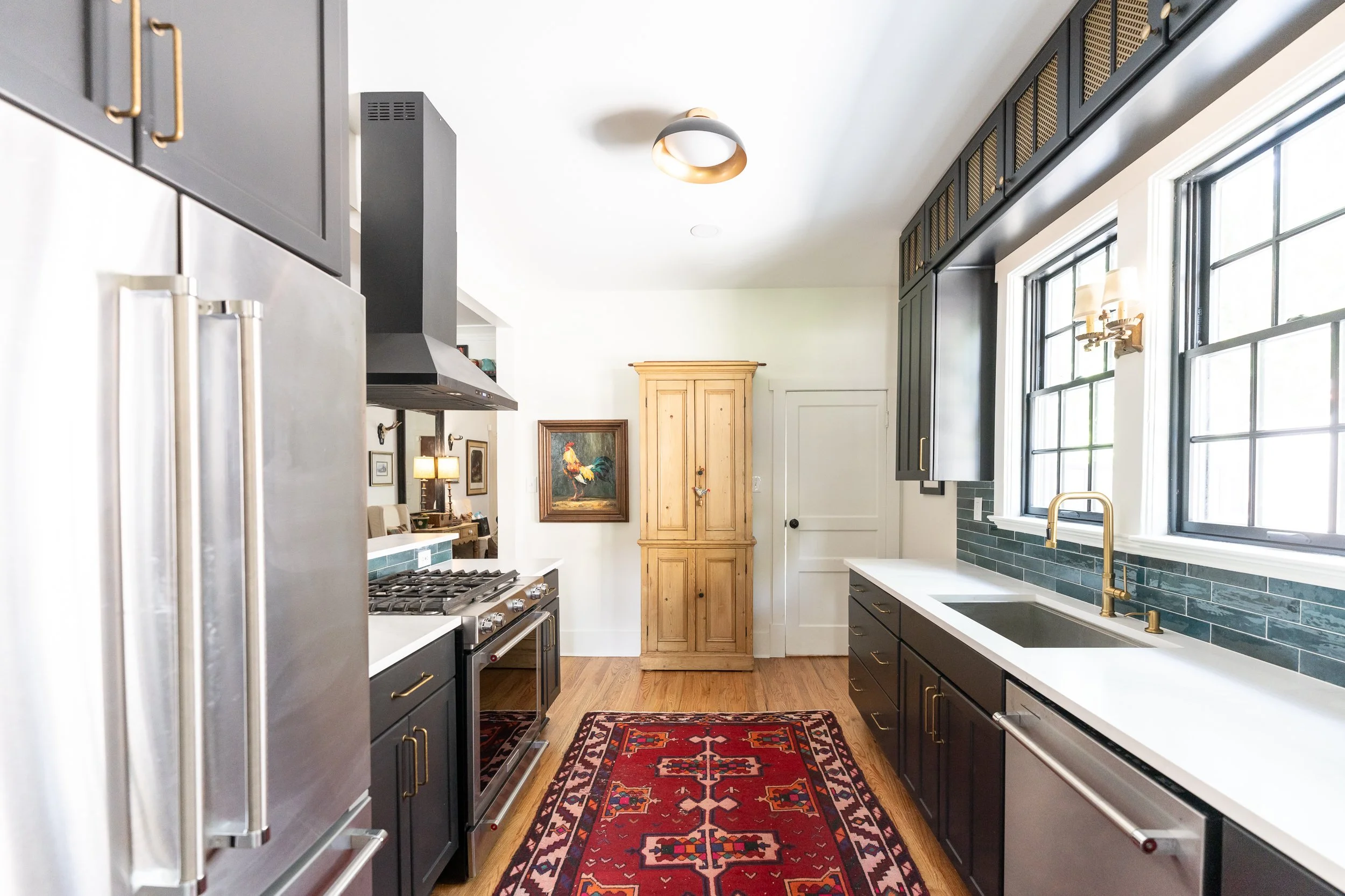A modern kitchen with black cabinets, stainless steel refrigerator, stainless steel oven, white countertops, and a window with black trim. There is a red patterned rug on the wooden floor, a wooden cabinet, and a colorful rooster painting on the wall.