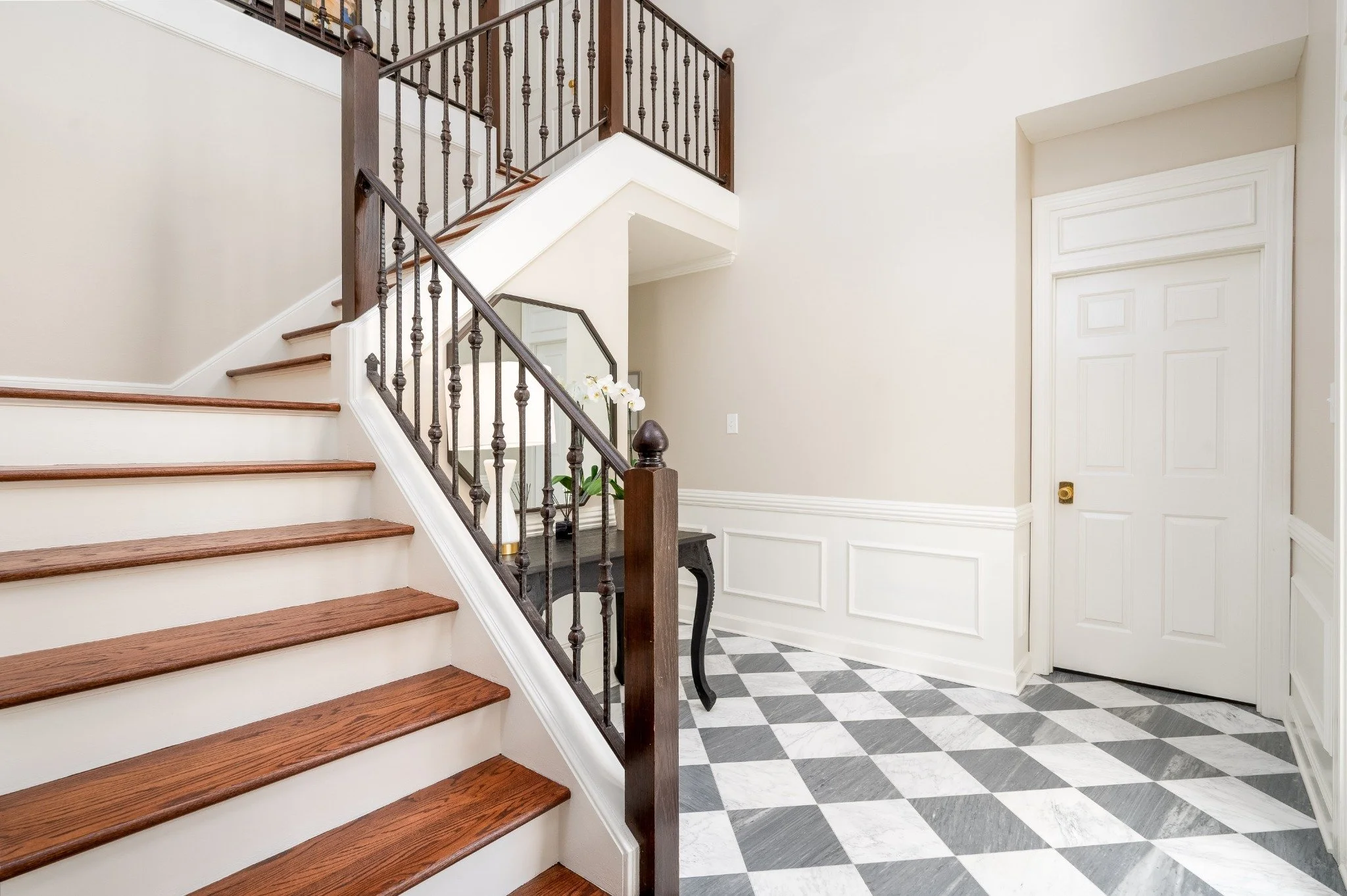 Interior view of a house entryway featuring a staircase with wooden steps, brass handrails, and black metal balusters. The floor has a black and white checkered tile pattern. White walls with decorative molding and a closed white door are visible.