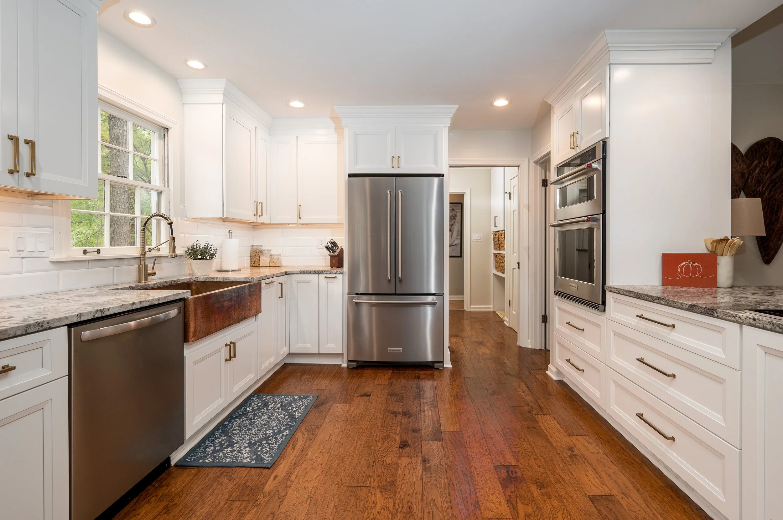 Bright white kitchen with wooden floors and white cabinets, stainless steel refrigerator, double ovens, large windows, granite countertops, and decorative items like a small plant and a framed pumpkin sign.