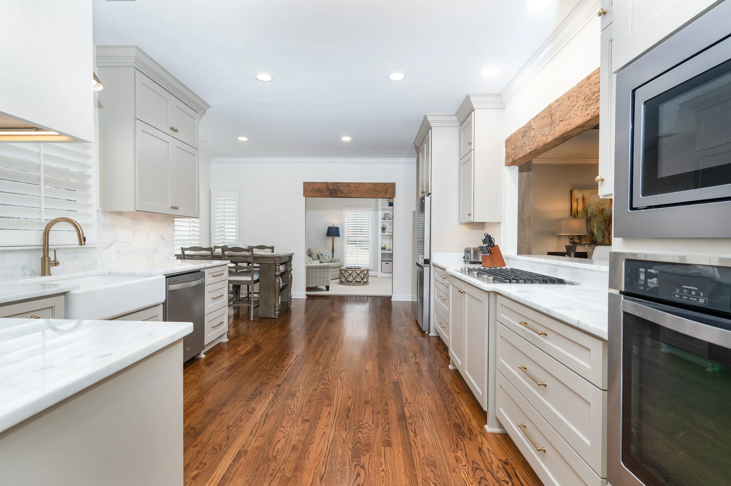 Spacious kitchen with white cabinets, marble countertops, stainless steel appliances, hardwood floors, and views into a living area with a dining table, chairs, and a cozy seating space with lamps and artwork.