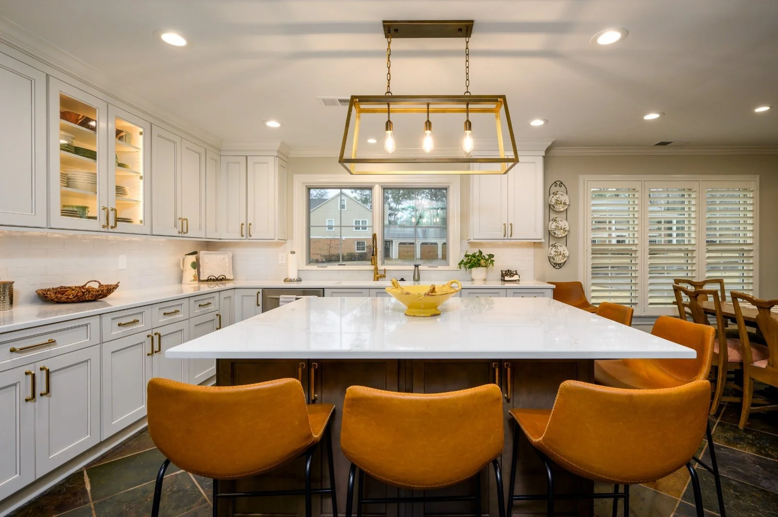 A modern kitchen with white cabinets, a central island with a white countertop, mustard-colored chairs around the island, and large windows with blinds, decorated with a pendant light fixture and some dishes on shelves.