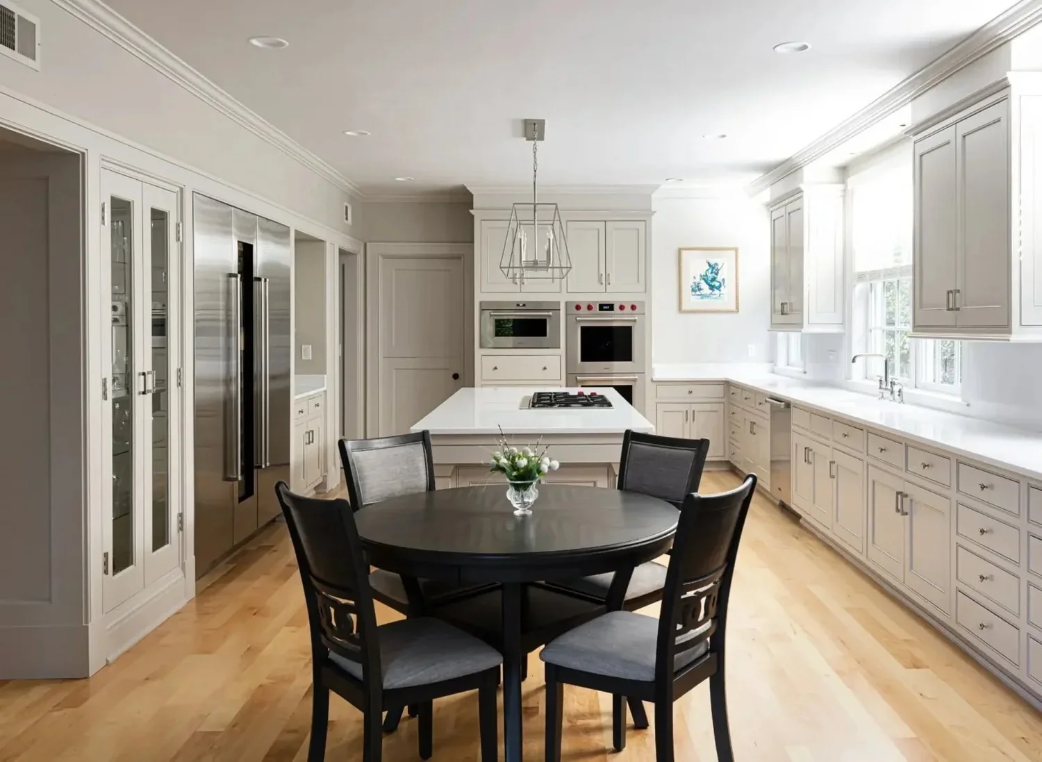Bright kitchen with white cabinets, a central island, a round black dining table with chairs, stainless steel refrigerator, built-in microwave and oven, and large windows providing natural light.