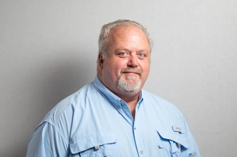 A middle-aged man with gray hair and beard wearing a light blue button-up shirt, standing against a plain light gray background.