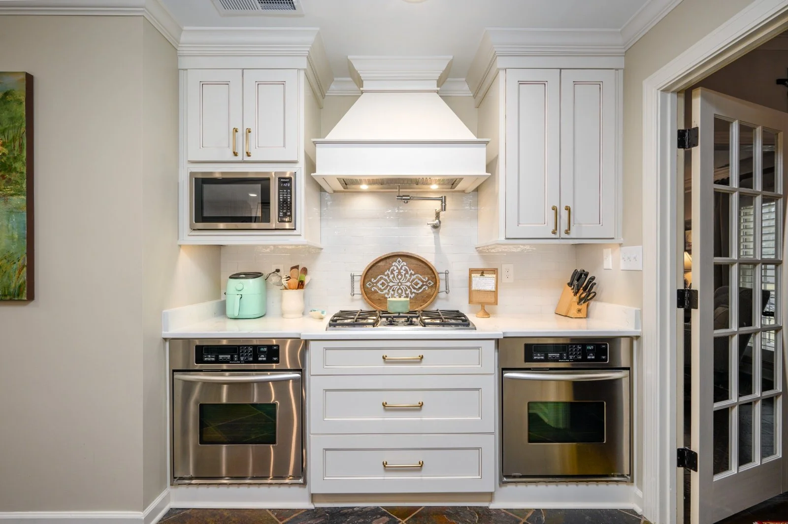 Kitchen with white cabinetry, stainless steel appliances, a gas stove, microwave, built-in ovens, and kitchen utensils on countertops.