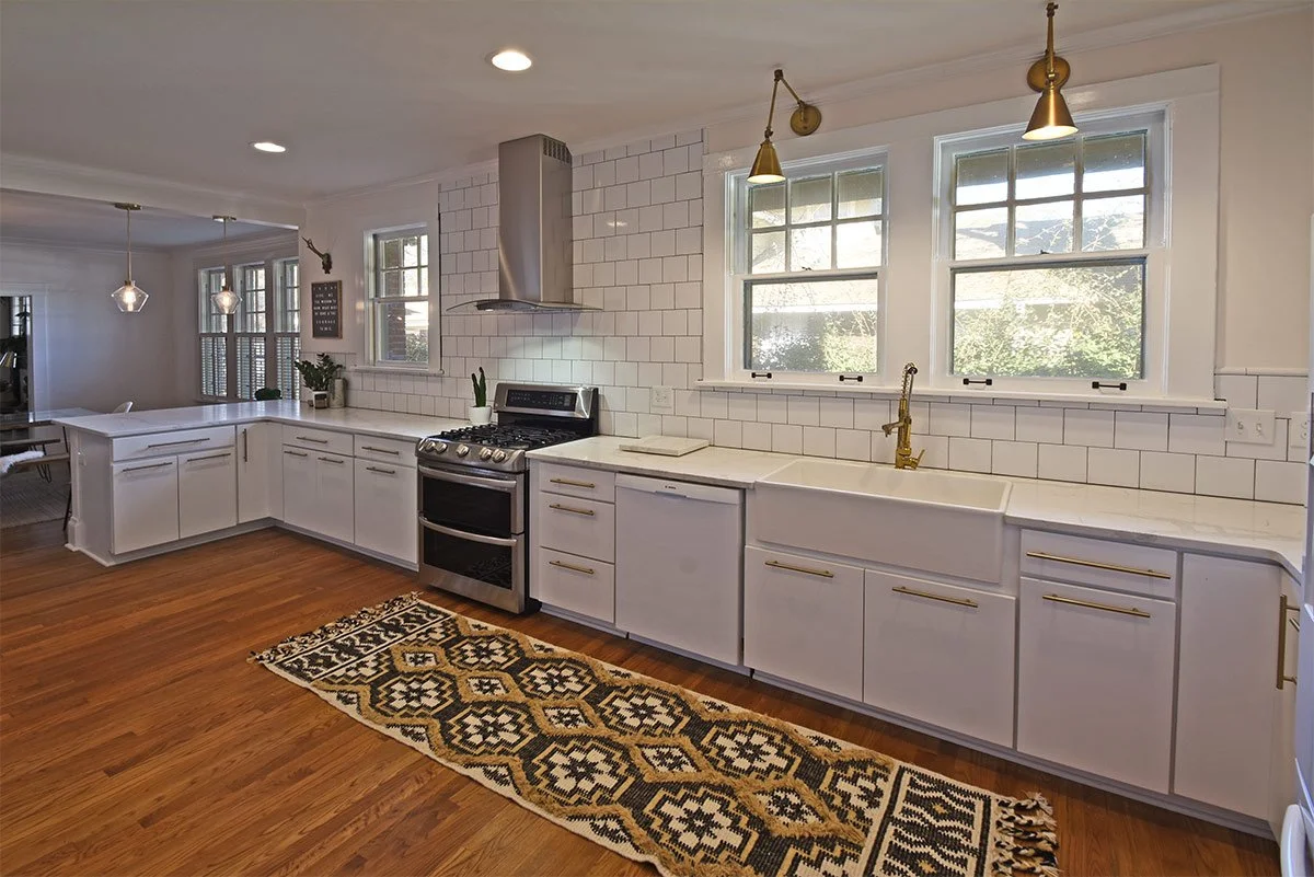 A bright kitchen with white cabinets, a white subway tile backsplash, and wooden floors. There is a stainless steel stove, a gold faucet over a large apron sink, and several windows letting in natural light. A patterned rug is on the floor in front of the sink.