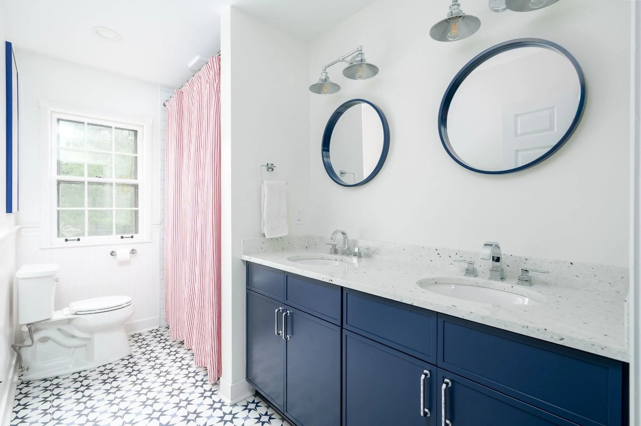 Upstairs bathroom remodel featuring a navy double vanity, white speckled countertop, round navy mirrors and patterned tile flooring.