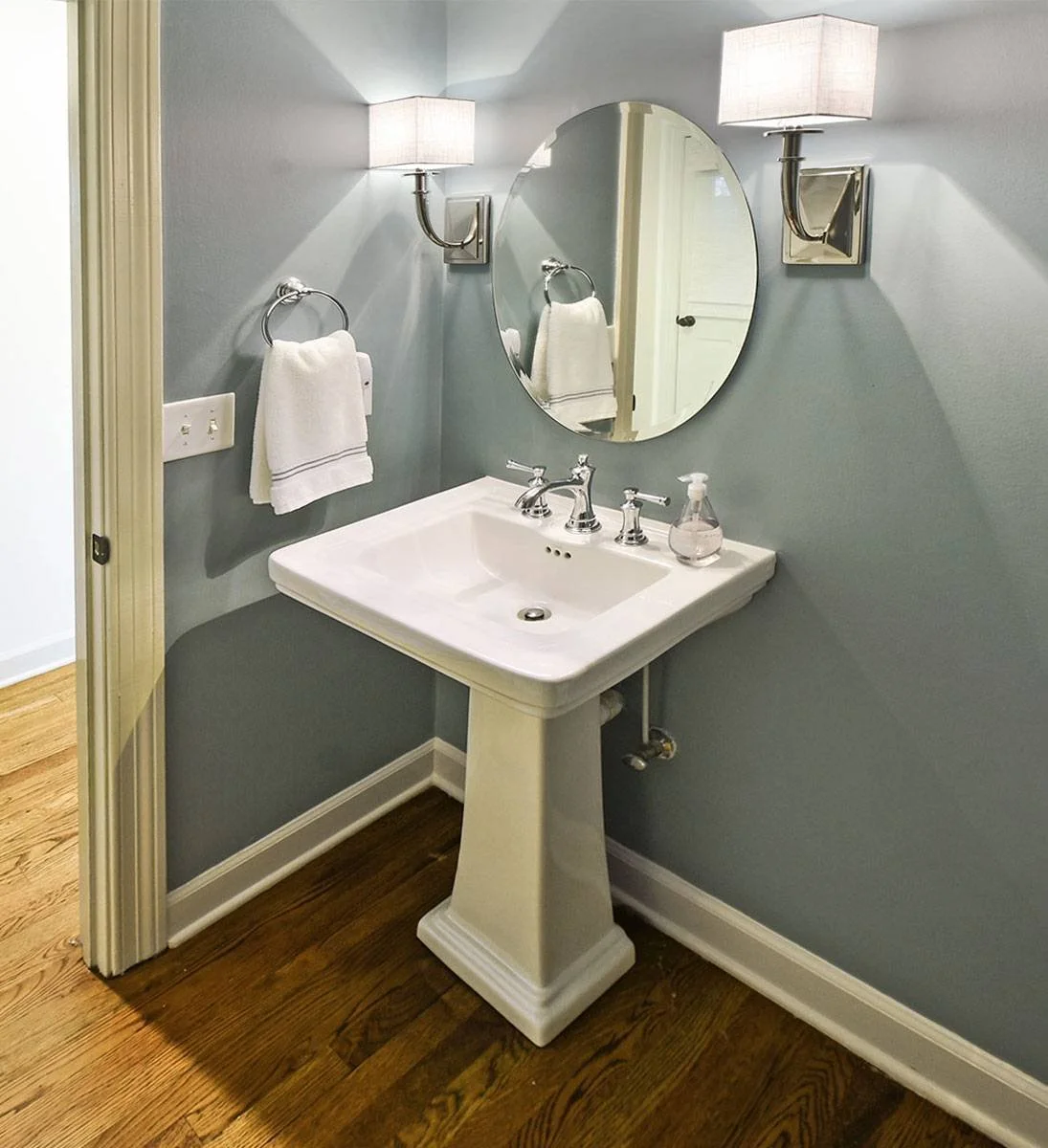 A clean and classic half bath featuring a pedestal sink, polished chrome fixtures, and soft blue-gray walls for a welcoming guest space.