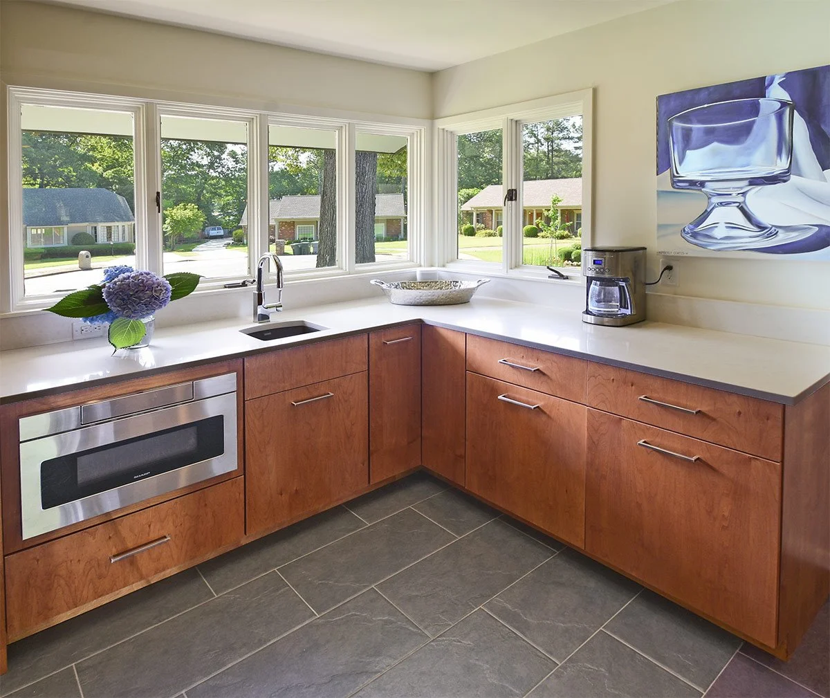 Kitchen with wooden cabinets, a white countertop, a window view of a suburban neighborhood, a bouquet of purple flowers, a bowl, a microwave, a coffee maker, and a wall art of a glass.