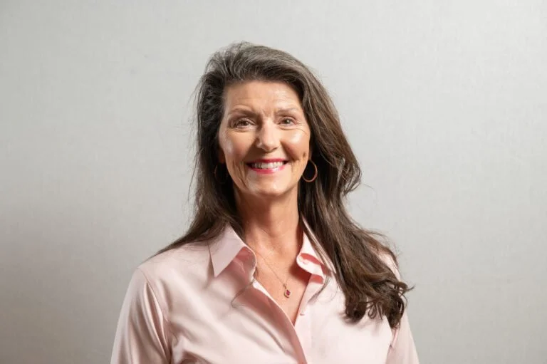 A smiling middle-aged woman with long, wavy brown hair, wearing a light pink button-up shirt and hoop earrings, standing against a plain, light-colored wall.