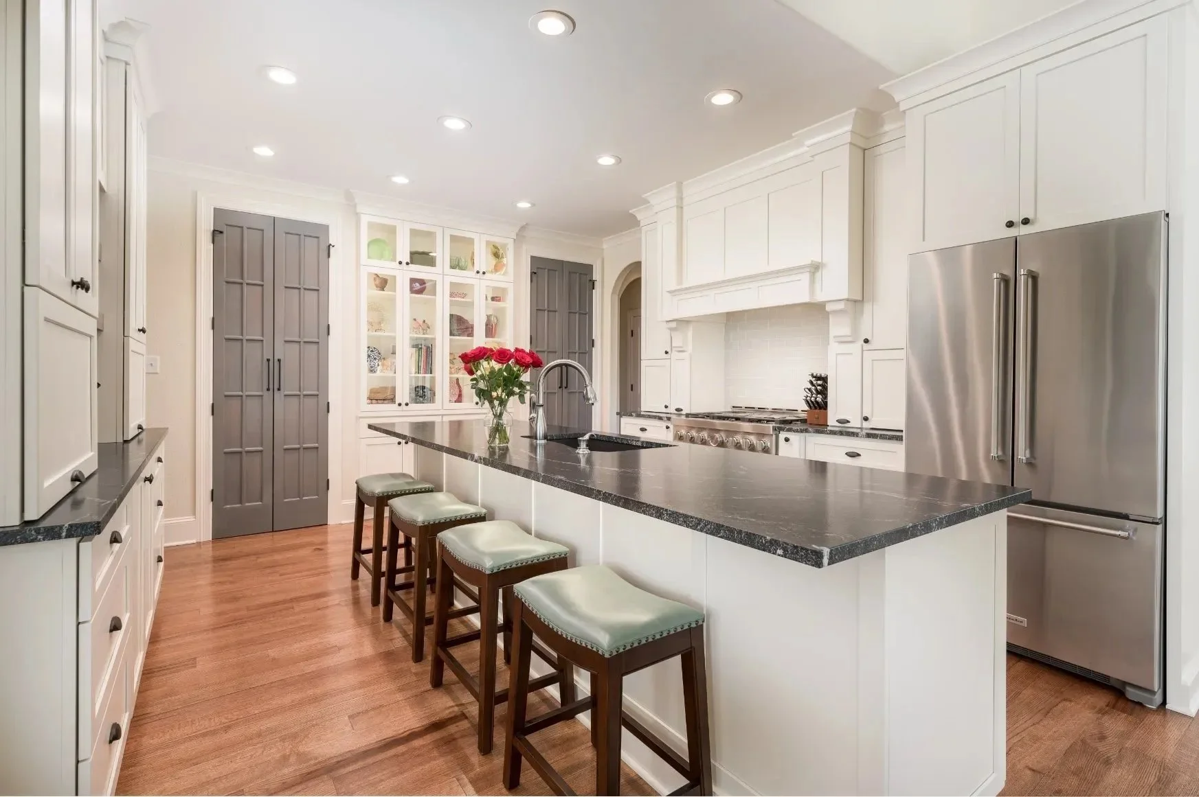 Modern kitchen with white cabinets, black granite countertops, stainless steel refrigerator, and a kitchen island with four barstools and a vase of red roses.