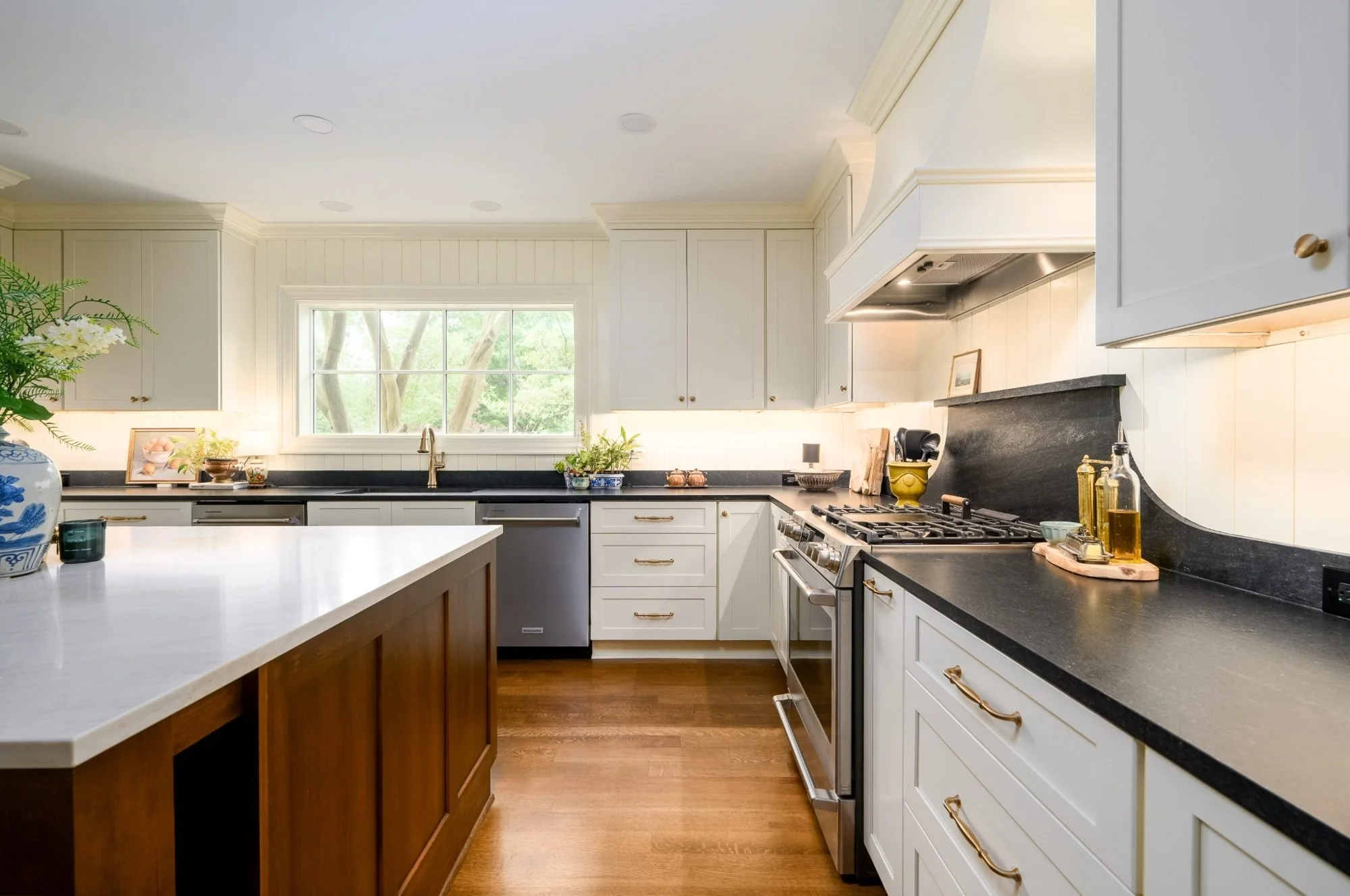 Bright custom kitchen remodel featuring white shaker cabinetry, black countertops, brass hardware, a professional-grade gas range, custom vent hood, and warm hardwood floors.