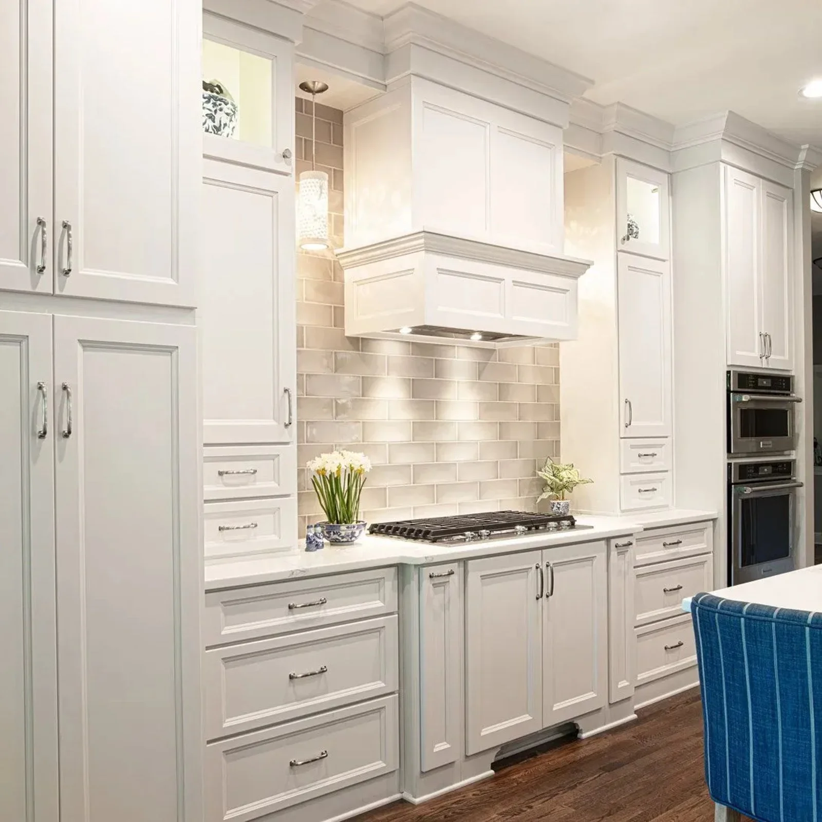 Custom kitchen remodel featuring light gray shaker cabinets, a decorative range hood, subway tile backsplash, quartz countertops, built-in double ovens, and rich hardwood flooring.