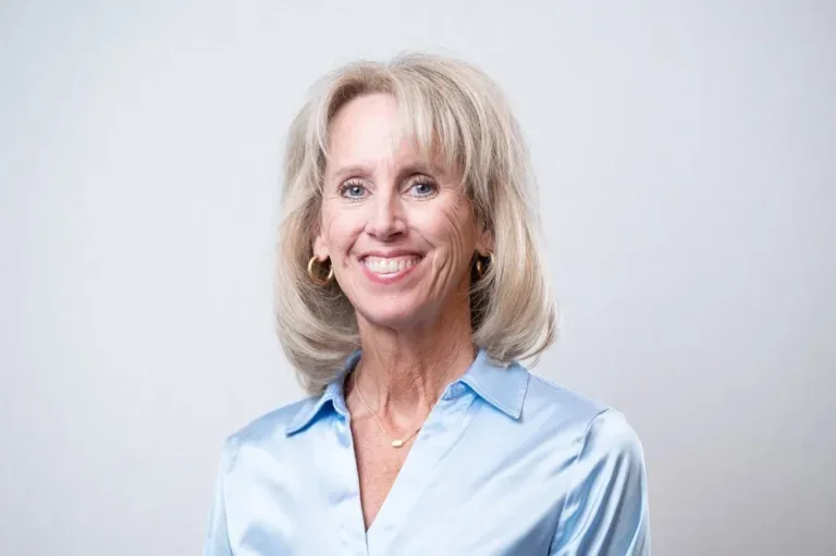 Portrait of a smiling woman with shoulder-length blonde hair, wearing a light blue button-up shirt, gold hoop earrings, and a gold necklace, standing against a plain light gray background.