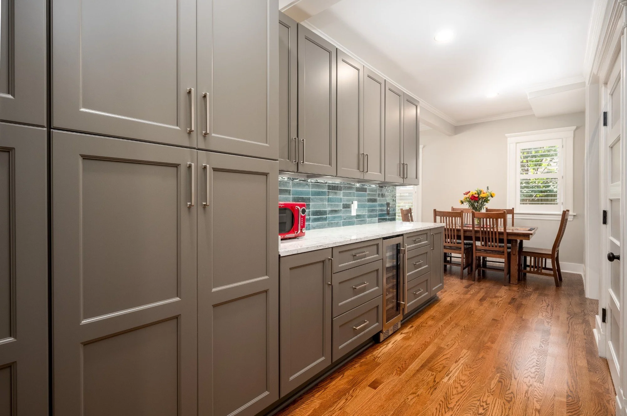 Interior view of a modern kitchen with gray cabinetry, a white marble countertop, a green tiled backsplash, and a wooden dining table with chairs. A window with blinds lets in natural light, and a vase with flowers decorates the table.
