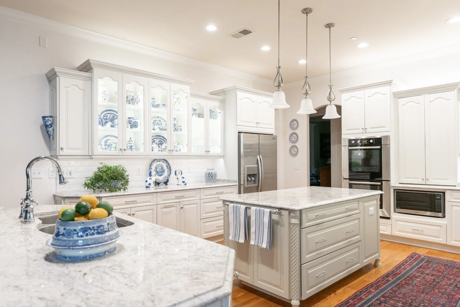 Bright white kitchen with granite countertops, white cabinetry, and a kitchen island. Decorated with blue and white dishware displayed in glass-front cabinets, a bowl of lemons and limes on the island, and a small green plant near the sink. Stainless steel refrigerator and oven, pendant lights, and a colorful rug on the wooden floor.