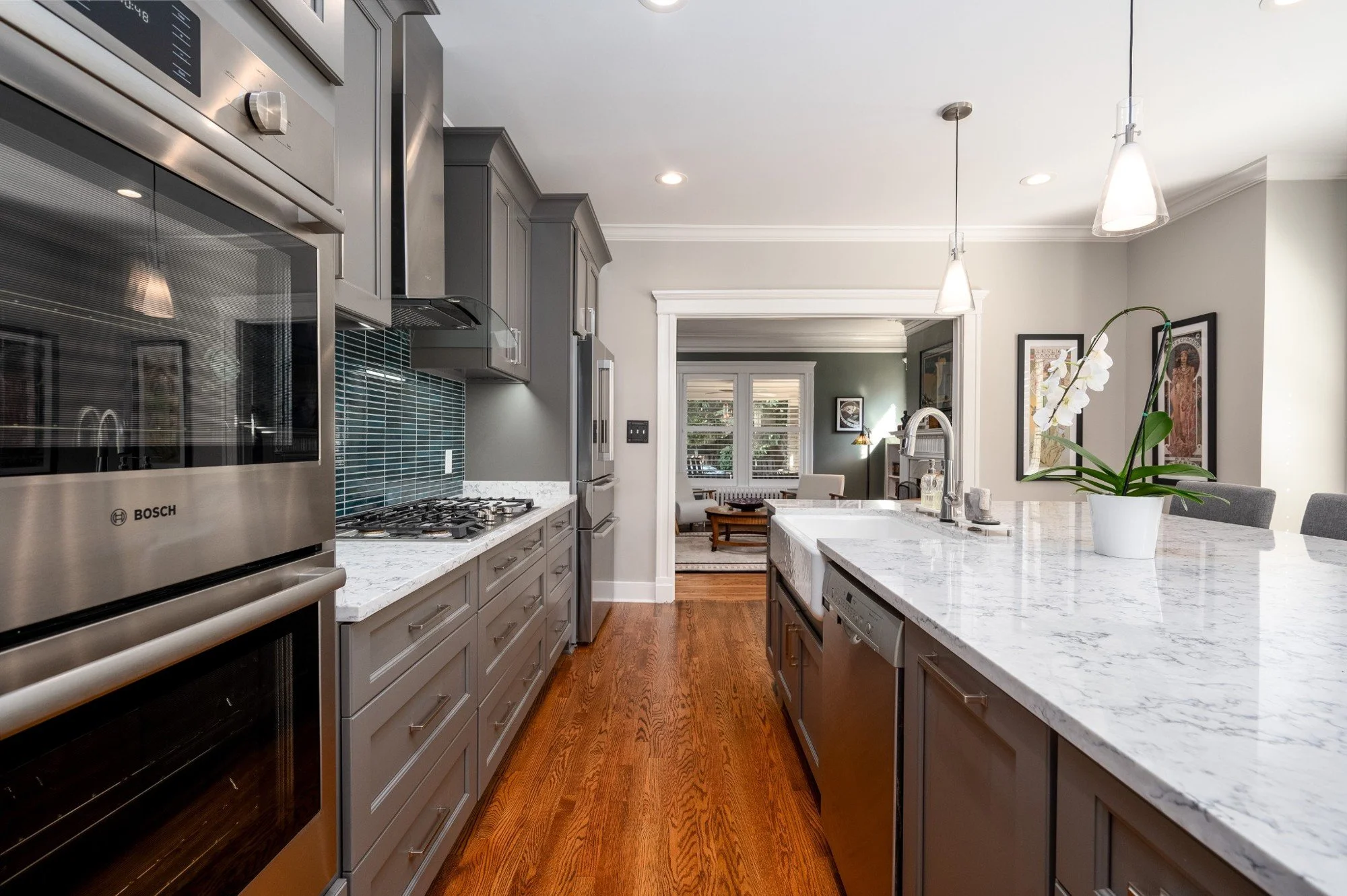 Modern kitchen featuring stainless steel appliances, gray cabinets, and a white marble countertop island with a potted orchid plant. The background shows an open living room with framed artwork and large windows.