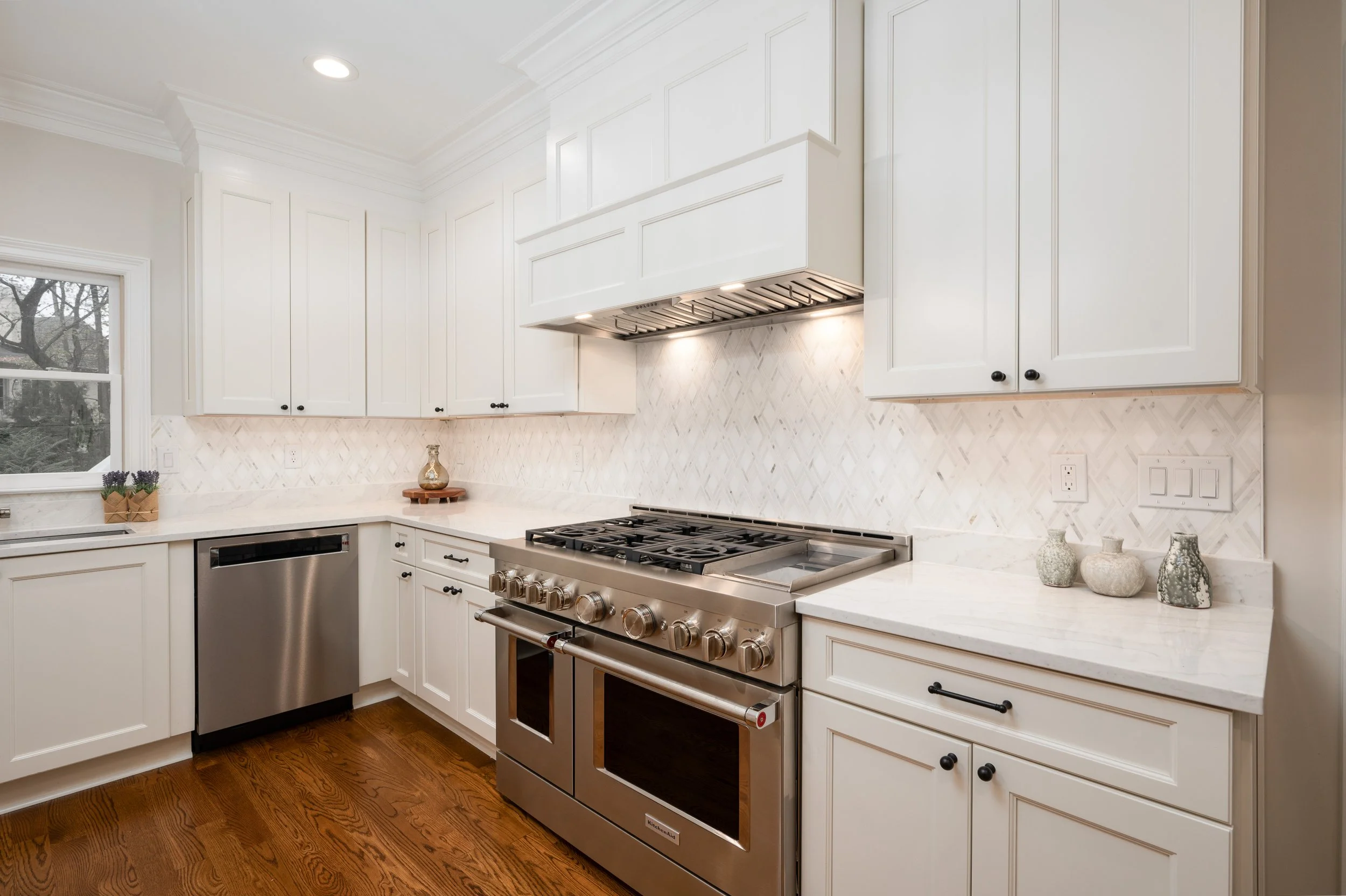 A modern kitchen with white cabinets, a stainless steel stove, and a marble countertop. There is a window with a view of trees outside, and decorative vases on the counter.