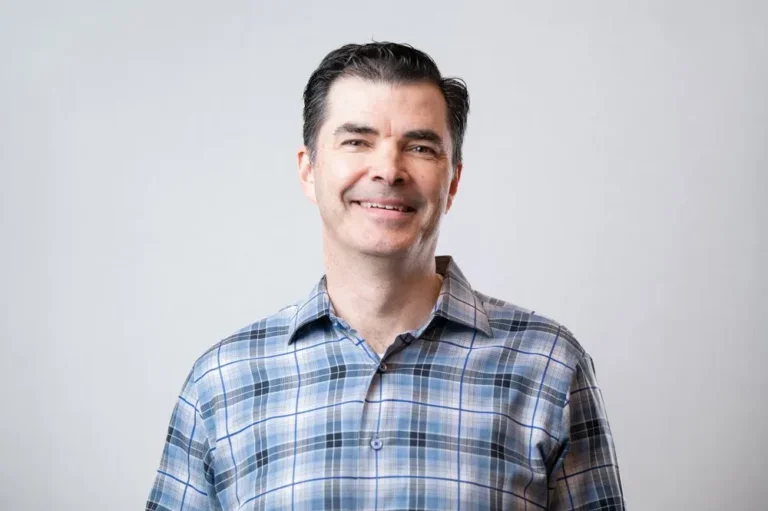 A smiling man with dark hair wearing a blue plaid shirt against a plain light gray background.