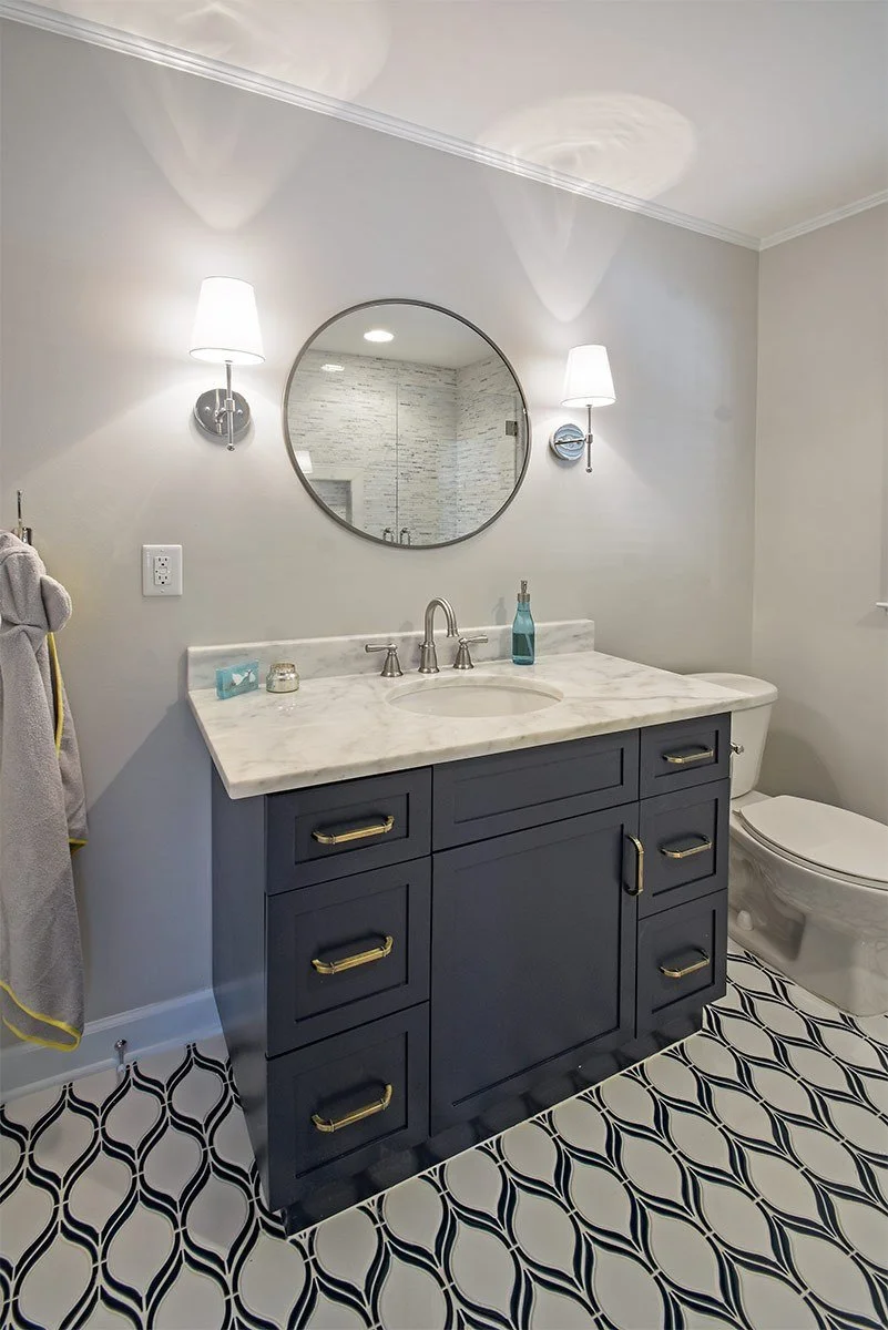 Bathroom with a navy blue vanity with gold handles, a marble countertop, an oval mirror, two wall-mounted white lamps, a decorative soap dispenser, and a toilet. The floor has black and white patterned tiles, and the wall is painted light gray.