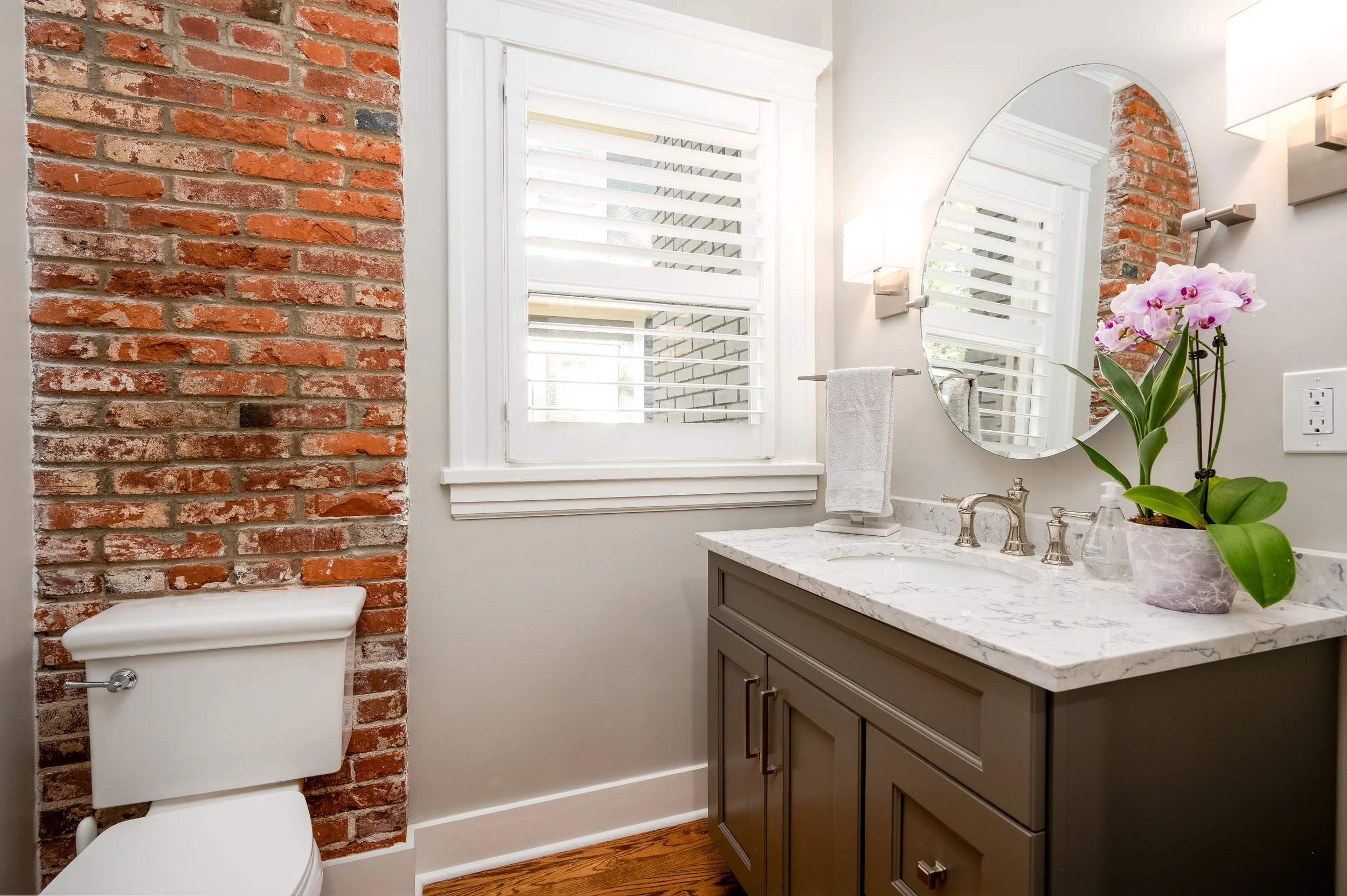 Bathroom with a brick accent wall, a window with white shutters, a wall-mounted mirror, a vase with pink orchids on a marble countertop, a towel bar with a white towel, and a toilet.