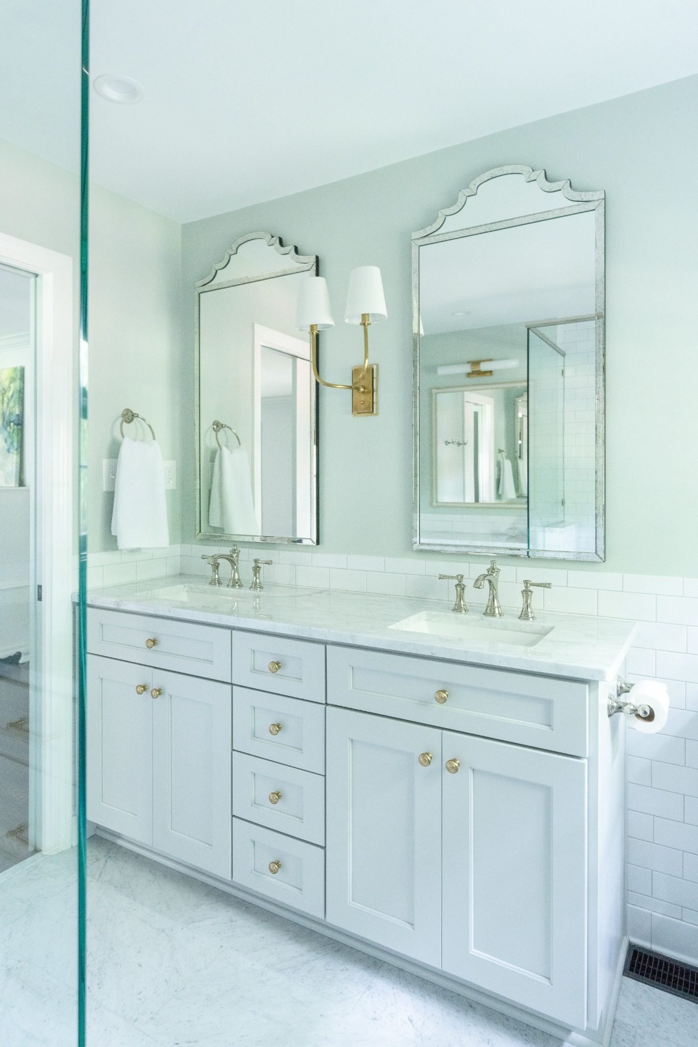 Bathroom vanity with white cabinetry, gold knobs, and two sinks with silver fixtures. Two mirrors above the sinks and a white wall-mounted light fixture with two white shades. Towels hanging on rings and a glass shower door on the left. Light-colored walls and white tiled backsplash.