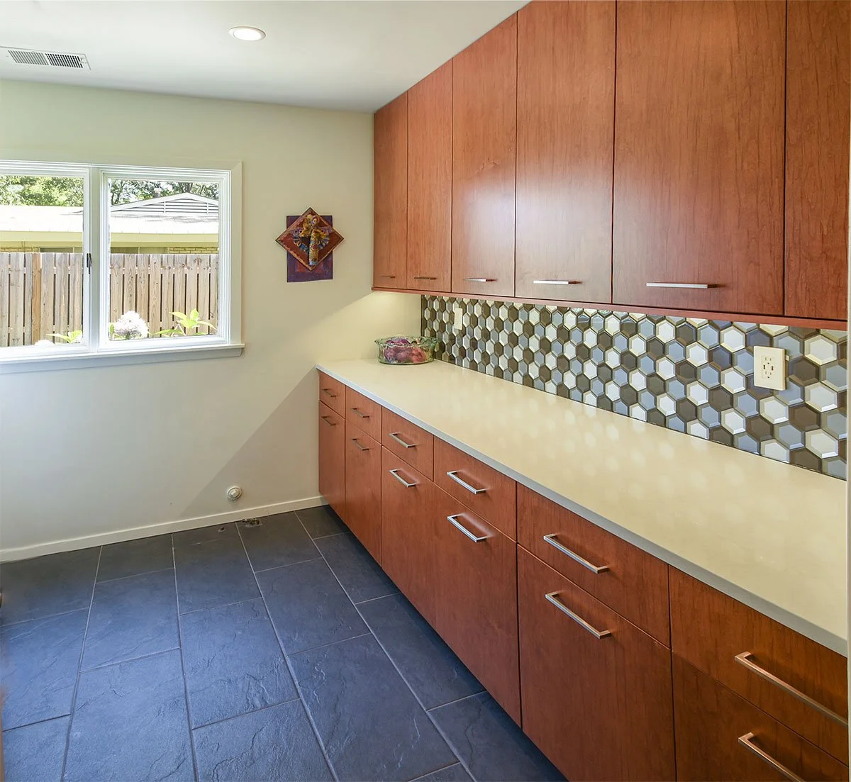 Kitchen with wooden cabinets, a yellow countertop, a honeycomb tile backsplash, and a window overlooking a wooden fence.