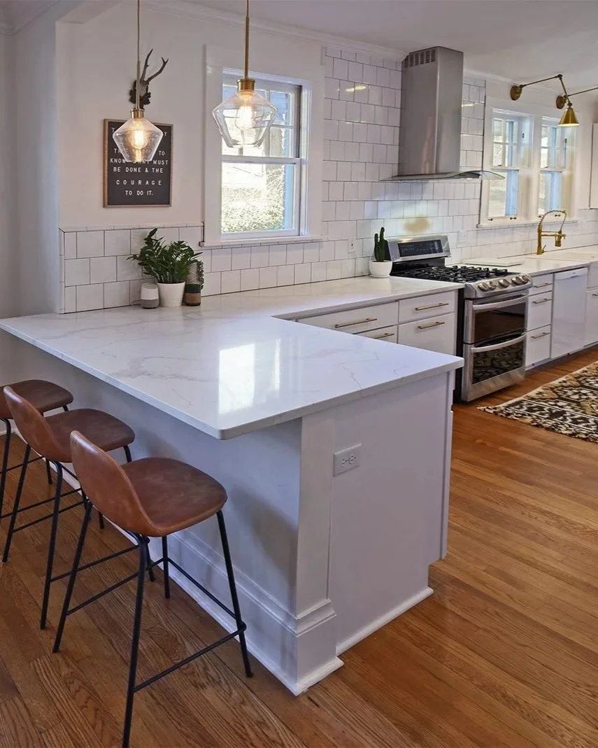 Modern kitchen with white cabinets, marble countertop, and hardwood floors. Features a stove, stainless steel range hood, white subway tile backsplash, and three pendant lights with geometric glass shades. Decor includes potted plants, a small framed sign, and a gold-brass faucet.