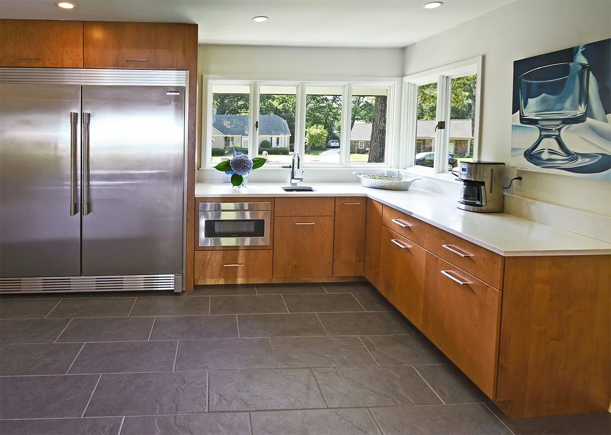 Kitchen with wooden cabinets, stainless steel refrigerator, white countertop, window with a view outside, coffee maker, and a painting of a glass on the wall.