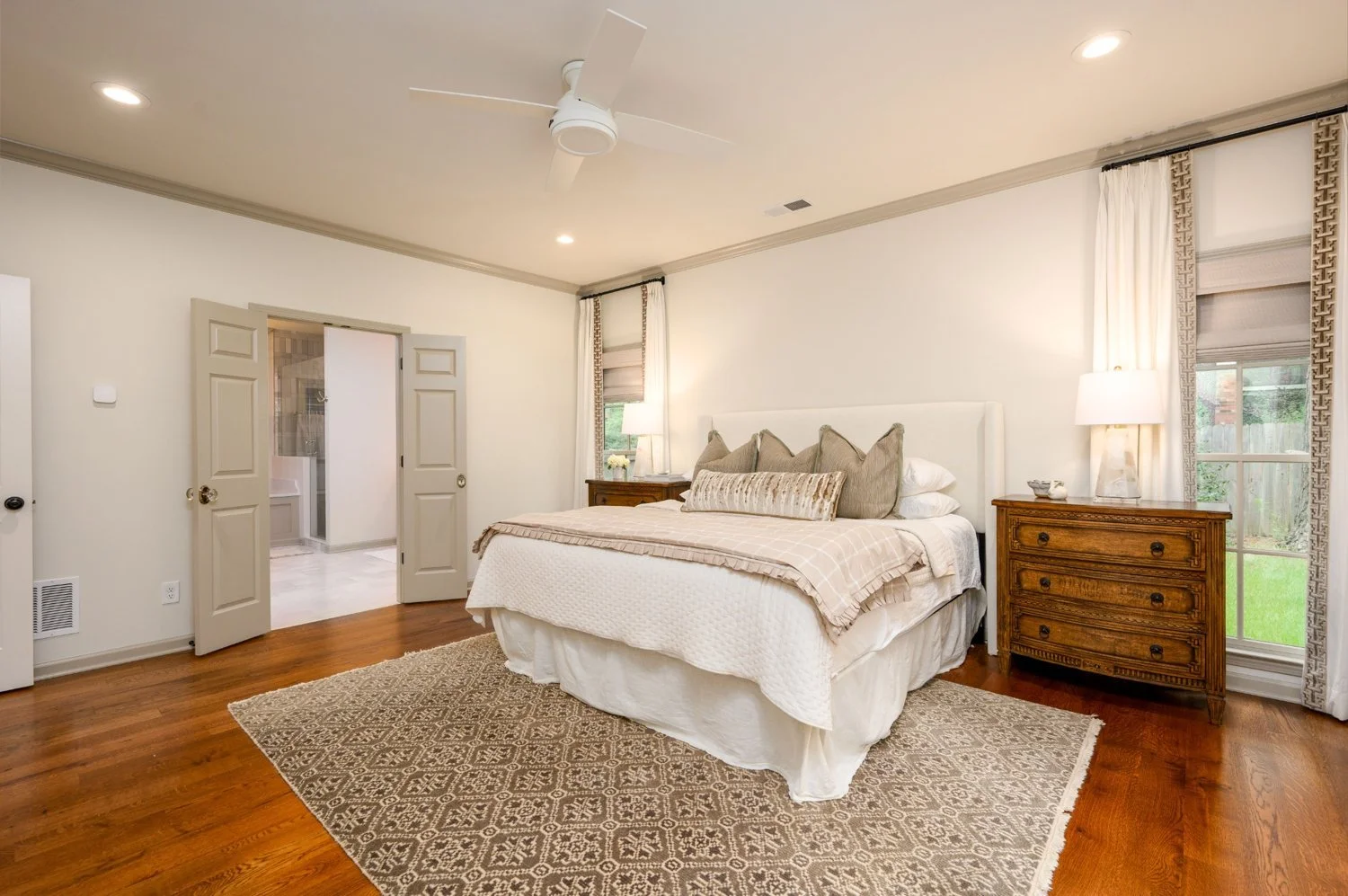 A cozy bedroom with a large bed dressed in neutral-colored bedding and multiple pillows. There are two wooden nightstands with lamps, a window with curtains on either side, and a door leading to an ensuite bathroom. The room has a ceiling fan, recessed lighting, hardwood floors, and a patterned area rug.