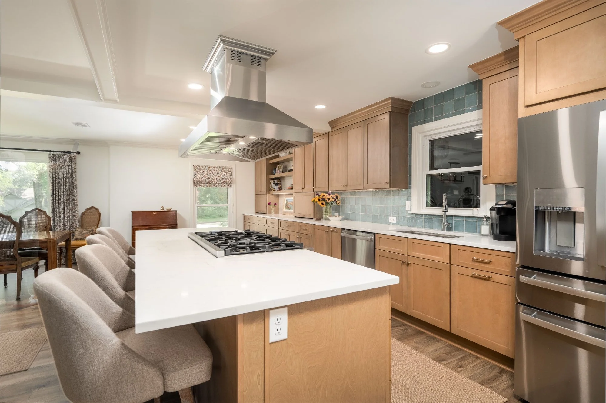 Kitchen with light wood cabinets, a white countertop island with beige upholstered chairs, a stainless steel range hood over a gas stove, and a window with a floral blind above the sink. There are appliances including a refrigerator and a coffee machine, and a vase of sunflowers on the counter.