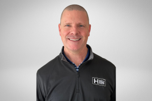 Portrait of a smiling man with short hair wearing a black jacket with a logo on the chest, standing against a plain white background.